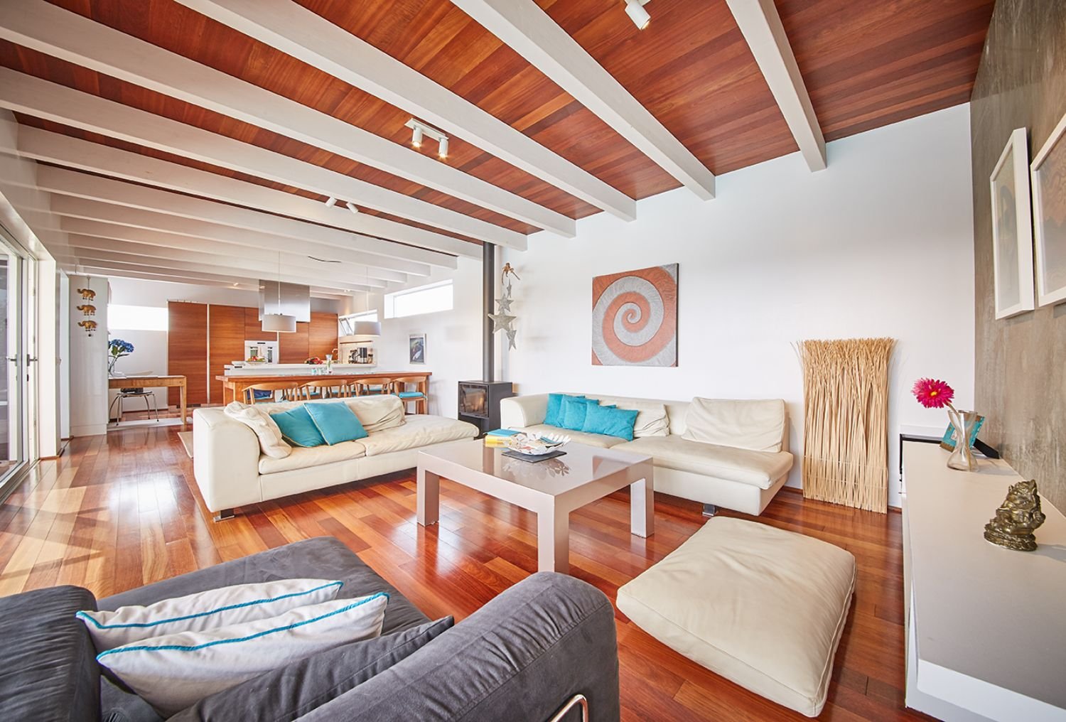 Moderne Wohnzimmer mit Holzboden und Holzbalkendecke, weißen Sofas und blauen Kissen, Couchtisch, Küchentisch im Hintergrund, Dekorationsgegenstände wie Statue und Gemälde.