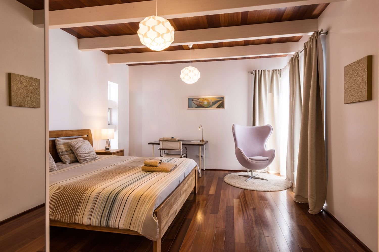 Modernes Schlafzimmer mit Holzfußboden, einem Bett, einem Sessel, einem Schreibtisch und Vorhängen.