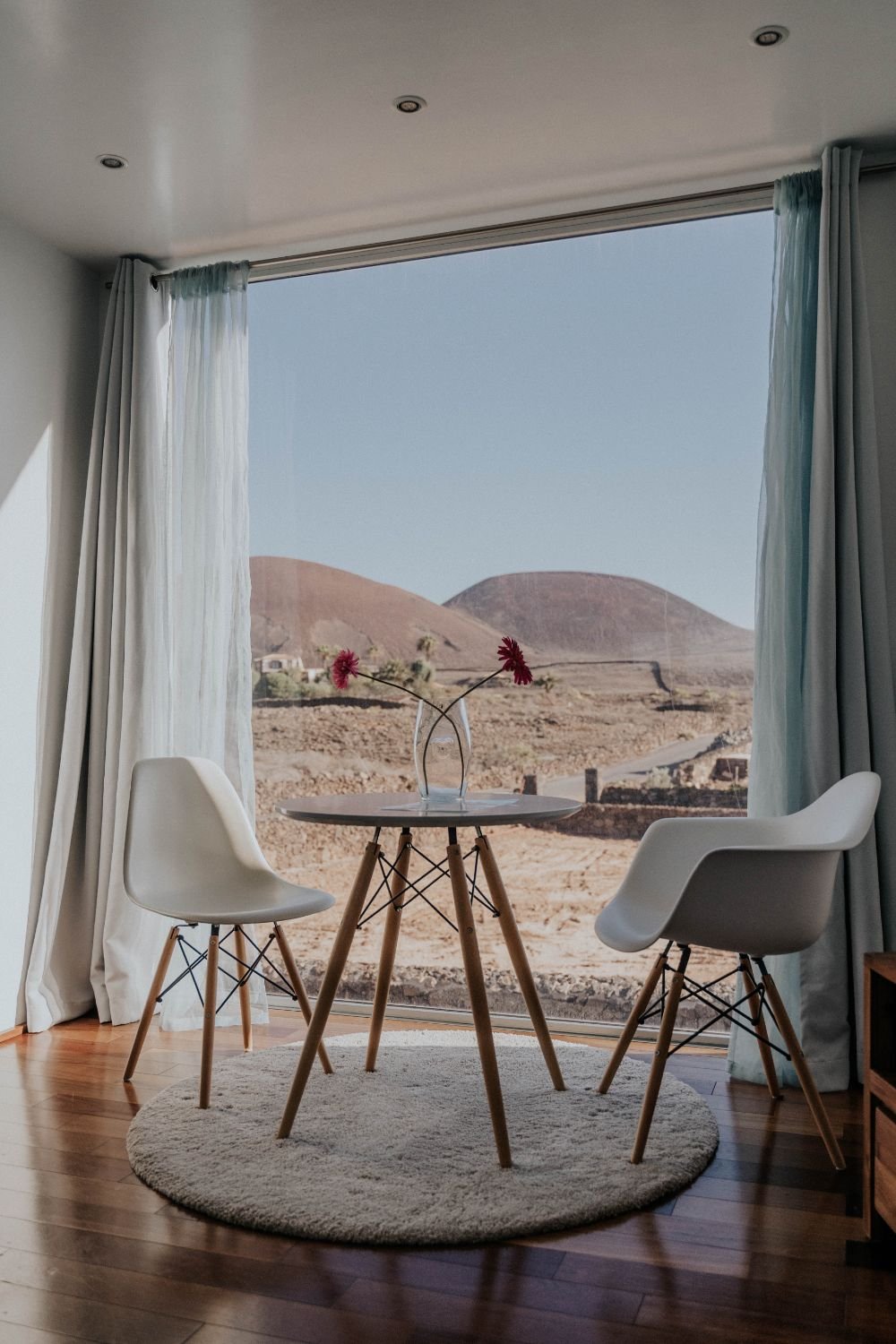 Innenraum mit zwei weißen Stühlen und einem runden Tisch, auf dem zwei Blumenvasen stehen, vor einem großen Fenster mit Blick auf eine Wüstenlandschaft und Hügel.