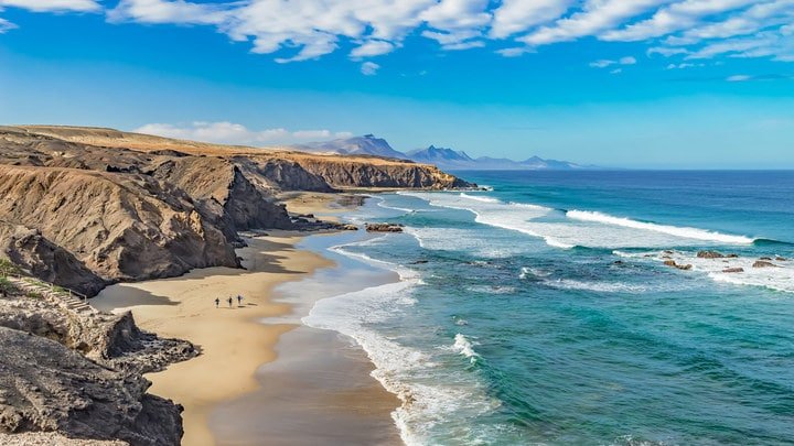Küstenlandschaft mit Klippen und Sandstrand, blauem Himmel und Wolken, Wellen des Ozeans und drei Personen, die am Strand spazieren.