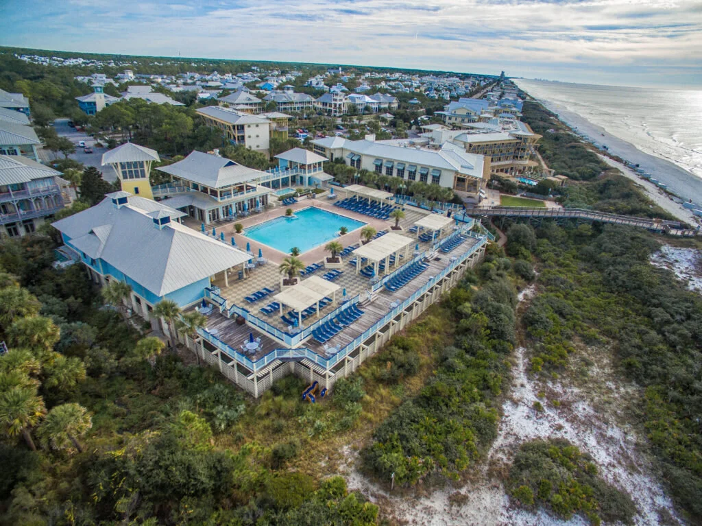 Aerial view of a beachside resort with a large swimming pool, surrounded by lounge chairs and umbrellas, with residential buildings and the ocean in the background.
