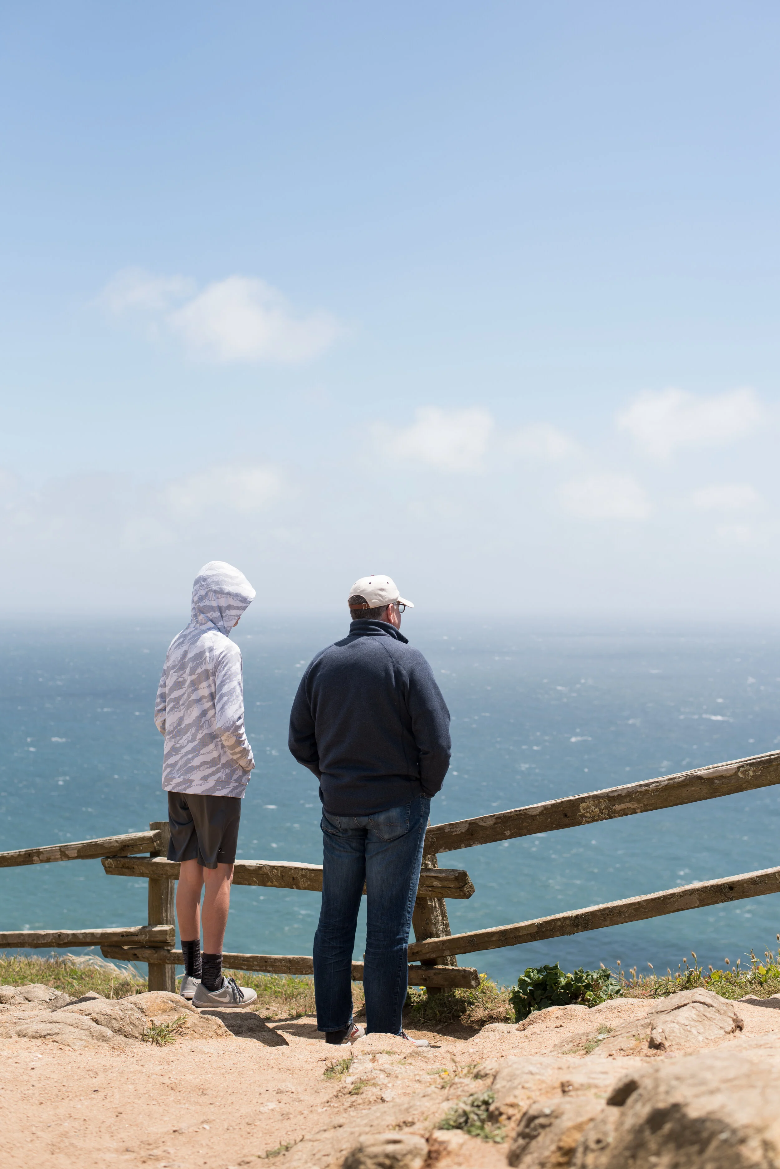 Three people standing on a rocky path near a wooden fence, overlooking the ocean under a partly cloudy sky. One person is wearing a hoodie, another in a jacket with a cap, and the third in a jacket with glasses.