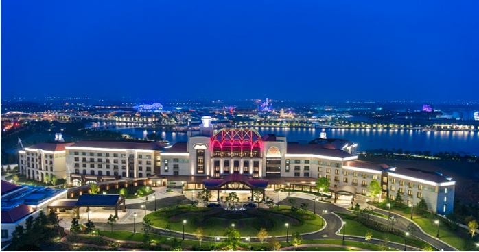 Nighttime aerial view of a large, illuminated hotel or resort with a central fountain, situated near a river with city lights in the background.