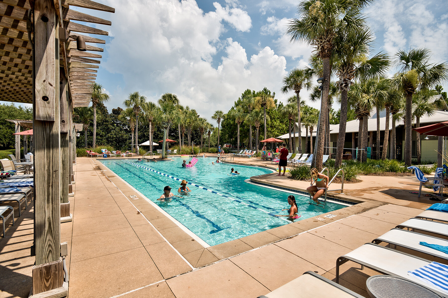 People swimming and relaxing at an outdoor pool surrounded by palm trees and lounge chairs on a sunny day.
