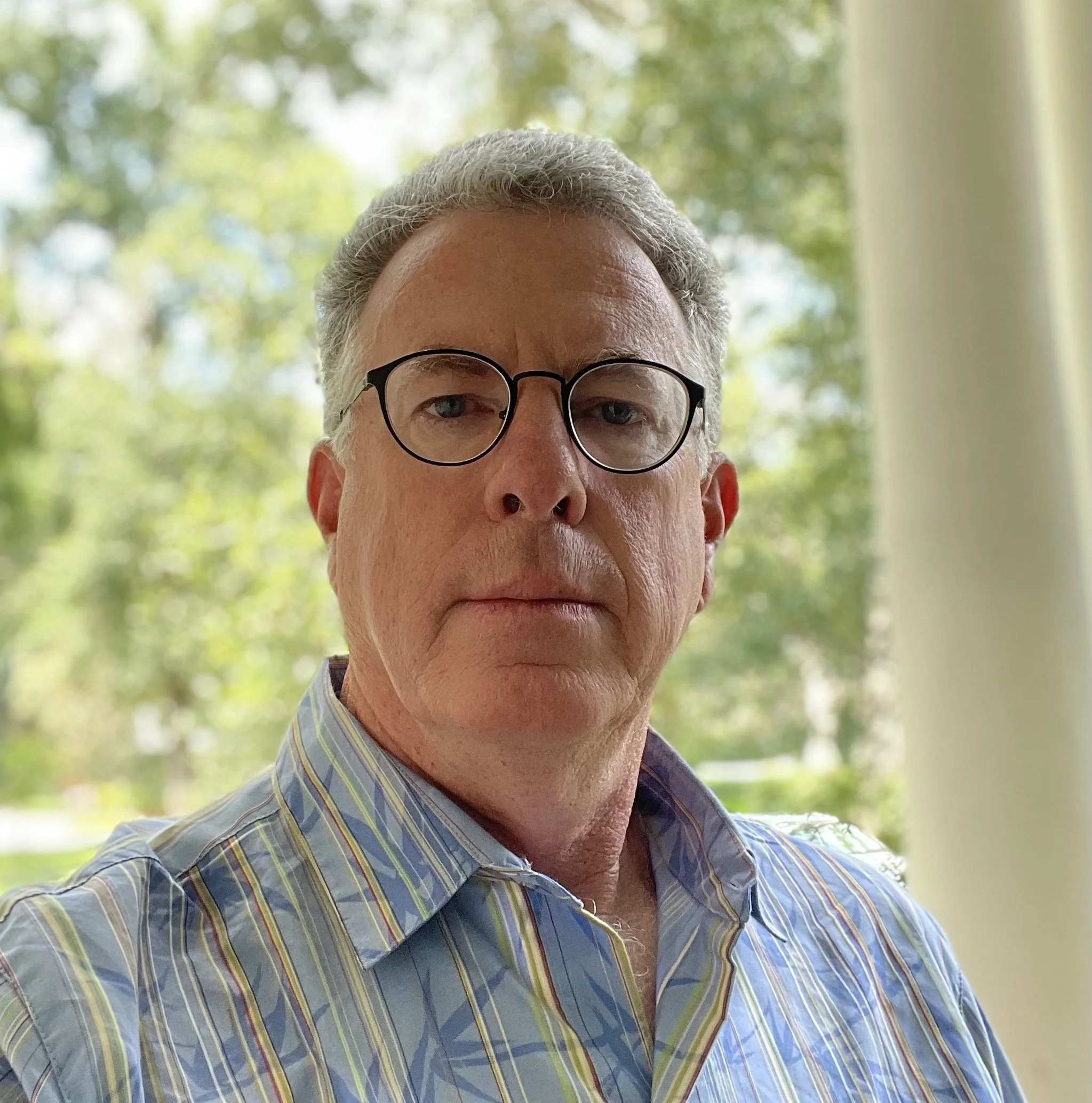 Close-up of a man's face with glasses, wearing a blue plaid shirt, outside near green trees.