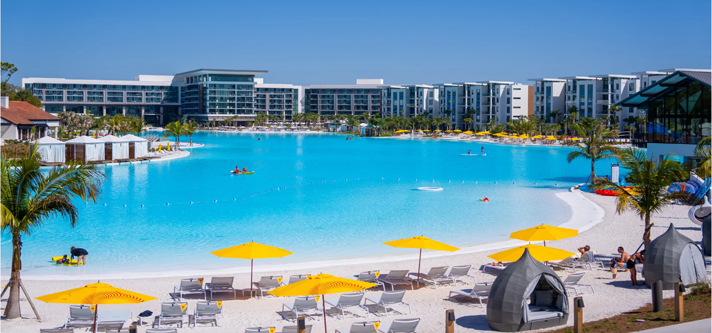 Resort with a large swimming pool, yellow umbrellas, lounge chairs, palm trees, and modern buildings in the background.