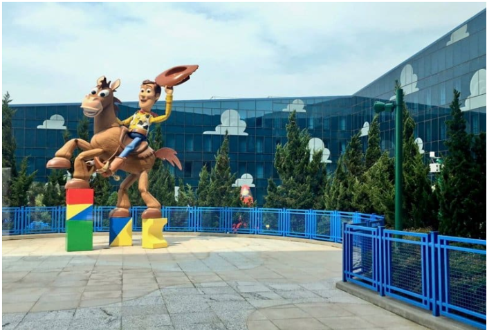 Statue of Woody Woodpecker riding a brown horse with a yellow saddle outside a modern building with cloud-shaped windows, surrounded by green trees and a blue fence.