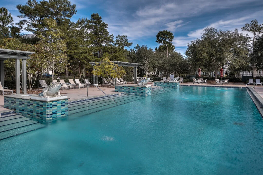 A serene outdoor swimming pool with crystal clear water, surrounded by lounge chairs, trees, and a partly cloudy sky.