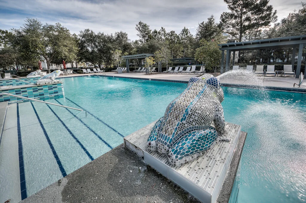 Empty outdoor swimming pool with mosaic frog sculpture on a pedestal, poolside lounge chairs, shaded seating area, and trees in the background