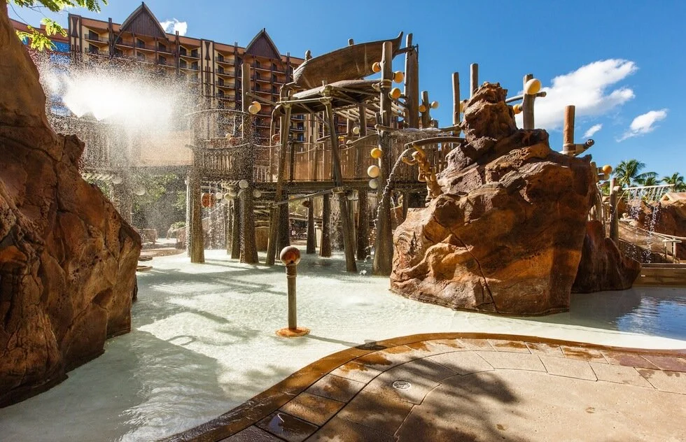 A water park with a wooden splash structure, rock formations, and water play features on a sunny day with blue skies.