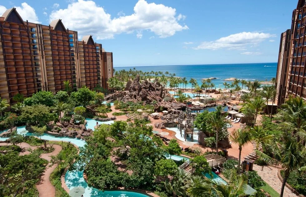View of a tropical resort with a large pool, lush greenery, palm trees, sandy beach, and ocean in the background under a partly cloudy sky.