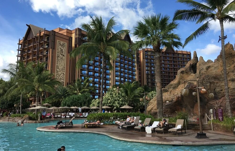 Resort with large swimming pool, palm trees, lounge chairs, and a tall hotel building in the background under a blue sky with clouds.
