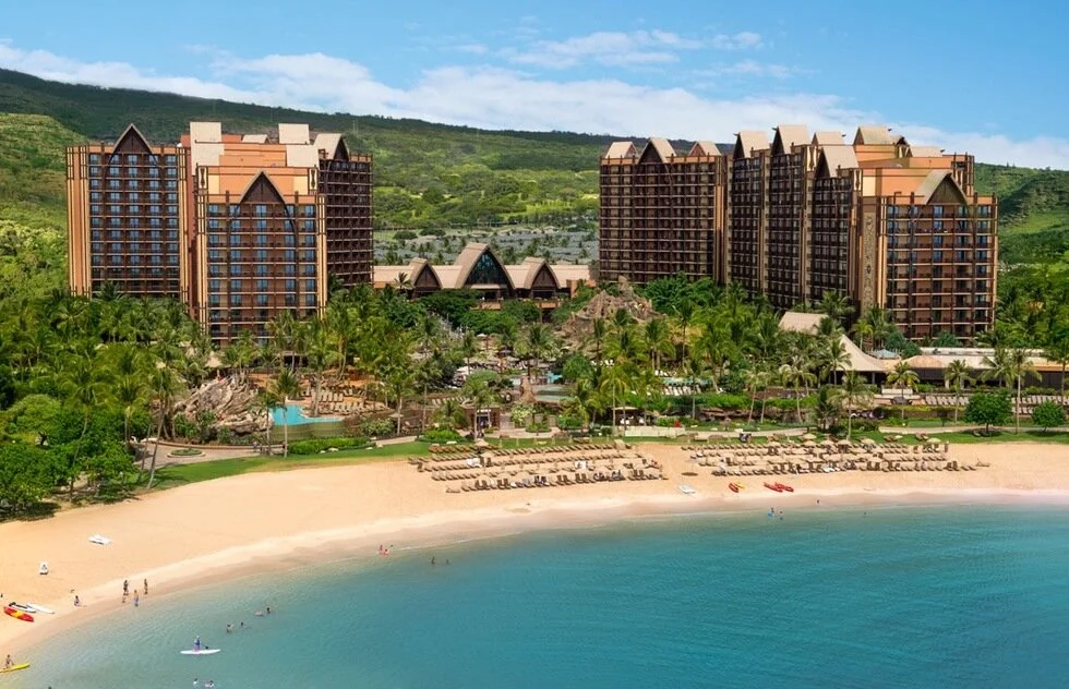Large beachfront resort with multiple buildings, swimming pools, palm trees, and a sandy beach with lounge chairs, facing clear blue water under a partly cloudy sky.
