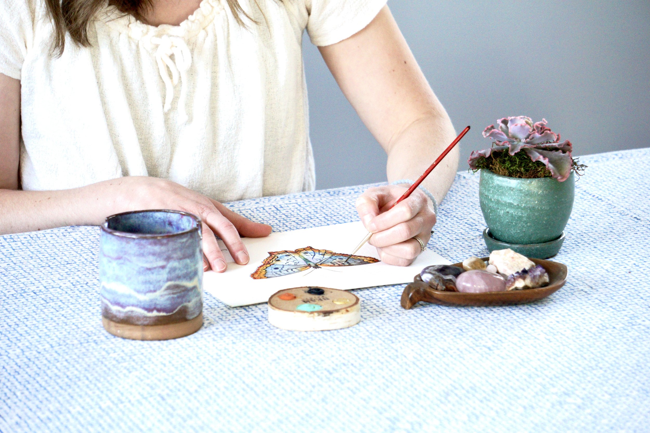 Person drawing a butterfly with watercolor paints on paper, seated at a table with a potted plant, a ceramic cup, and a bowl of stones and crystals.