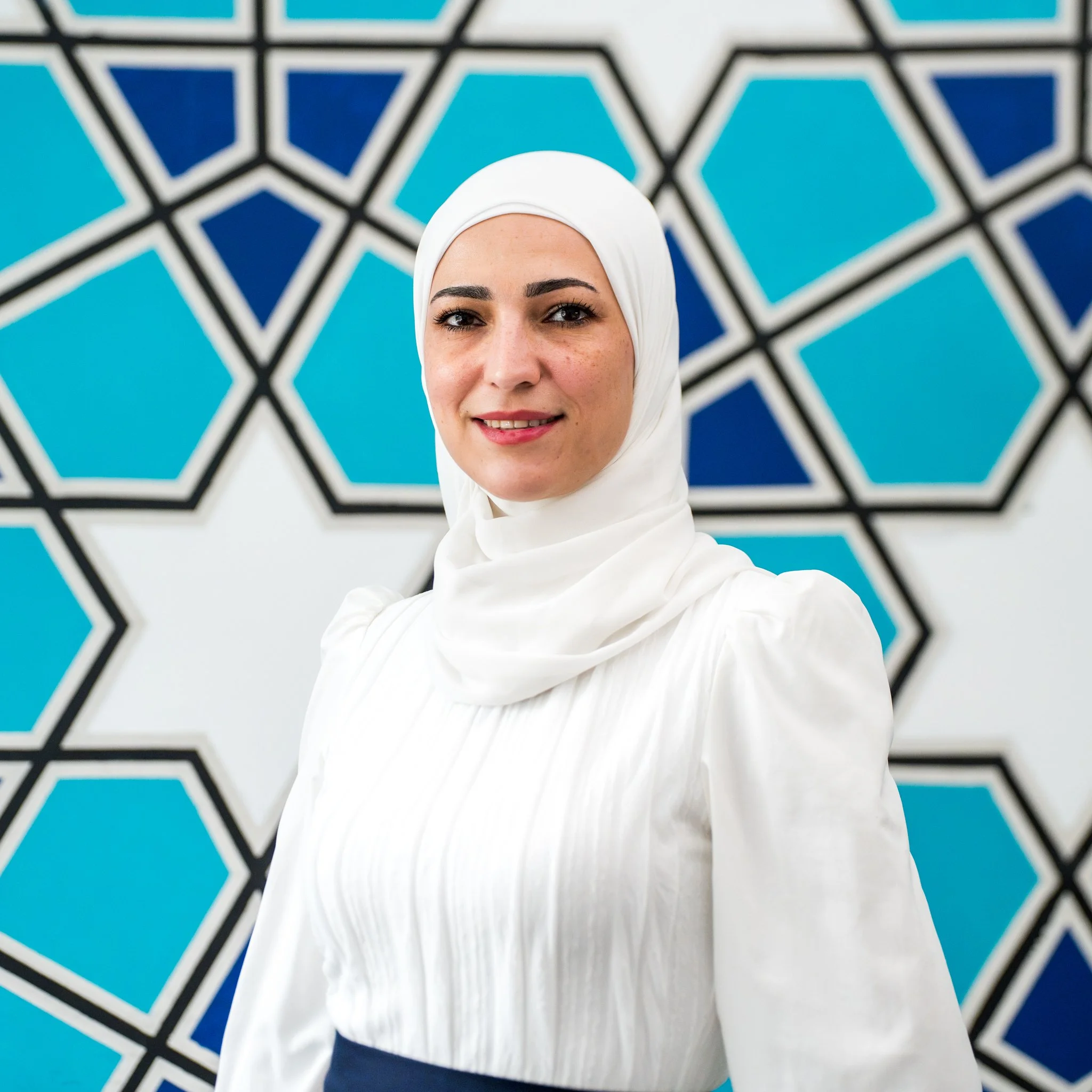 A woman wearing a white hijab and polka dot top smiling outdoors with trees and shaded structures in the background.