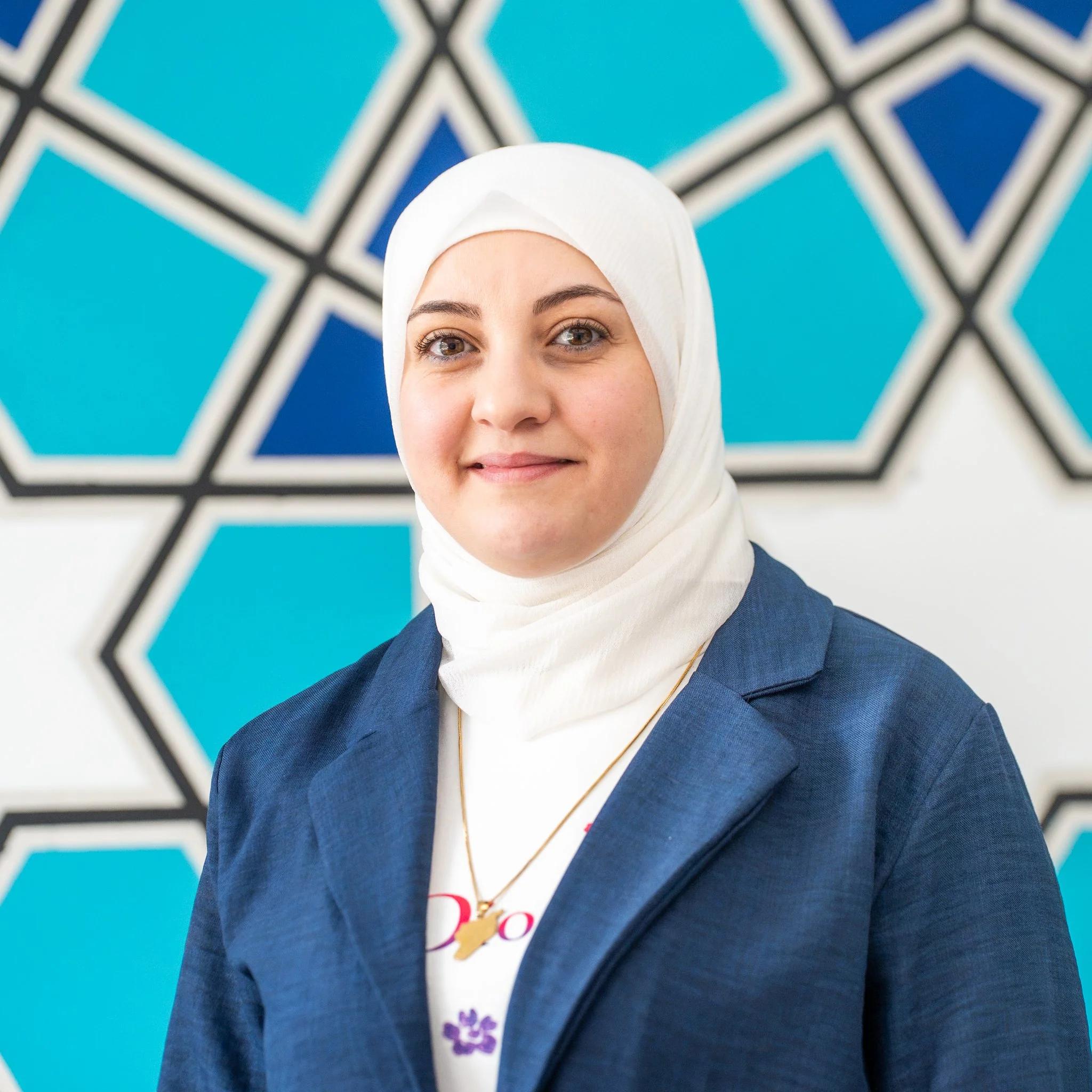 Smiling woman wearing a white hijab with pink accents, sunglasses on her head, and a blue denim jacket, standing outdoors near water and greenery.