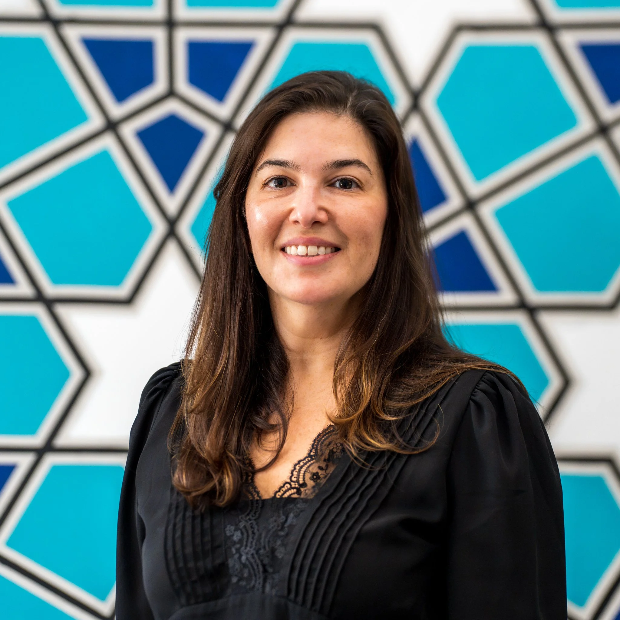 A woman with shoulder-length dark hair smiling, wearing a black dress with a floral pattern, standing in front of a gray backdrop with colorful circles and text.