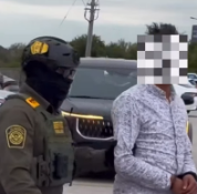 An ICE officer and detaining a person in a patterned shirt.