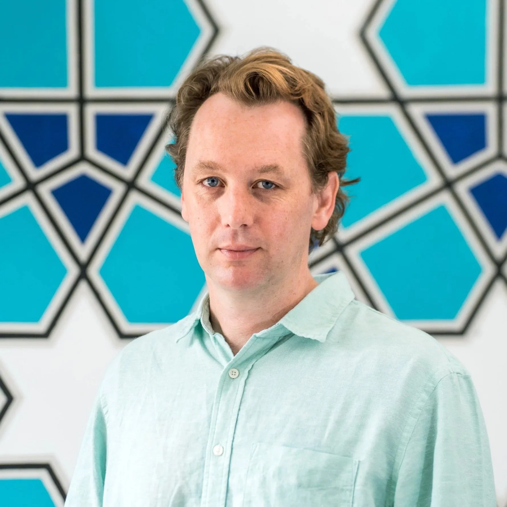 A man with light skin, brown hair, and blue eyes wearing a light blue button-up shirt, standing against a plain white wall.