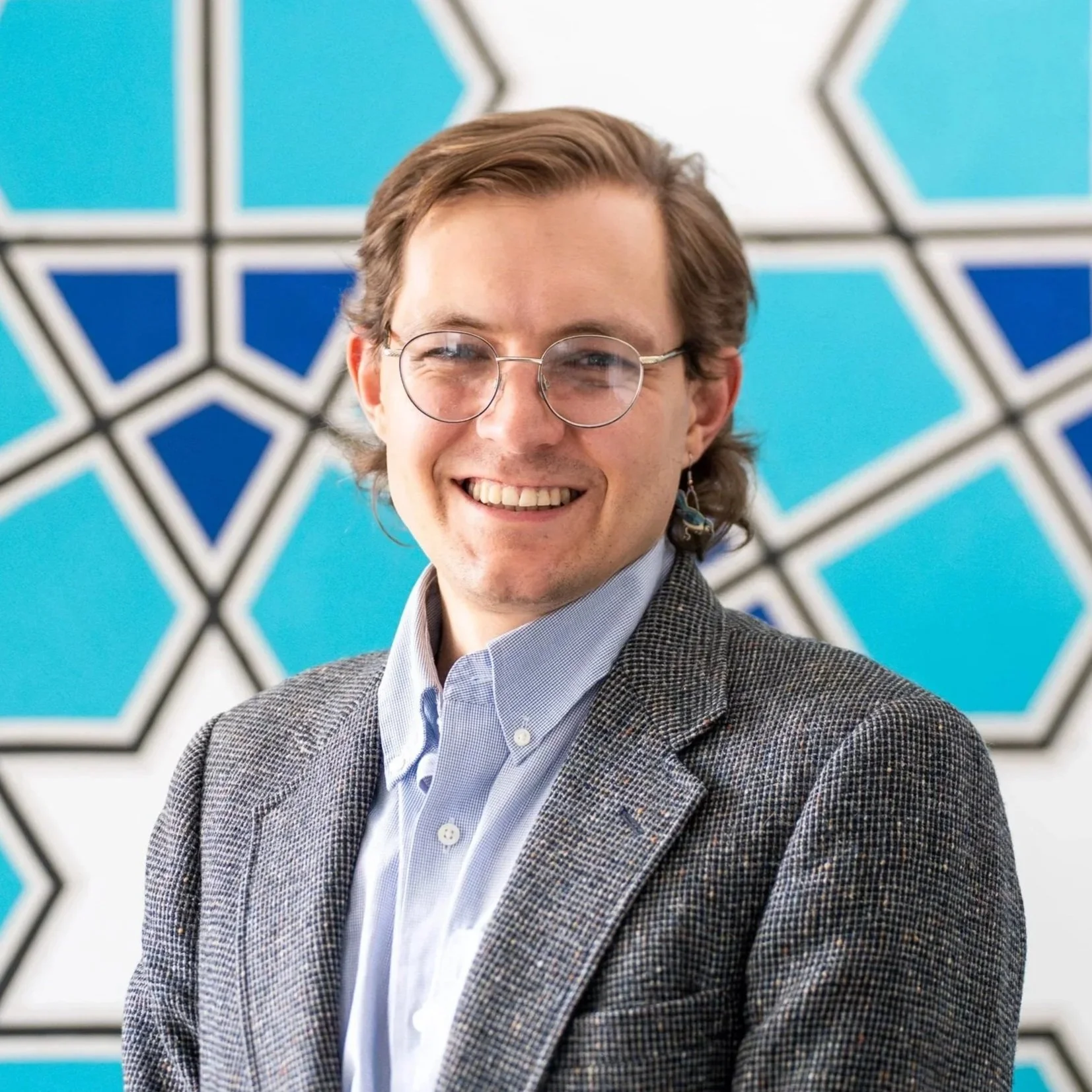 A person with short wavy hair wearing glasses and a white button-up shirt smiling at the camera, standing against a plain background.