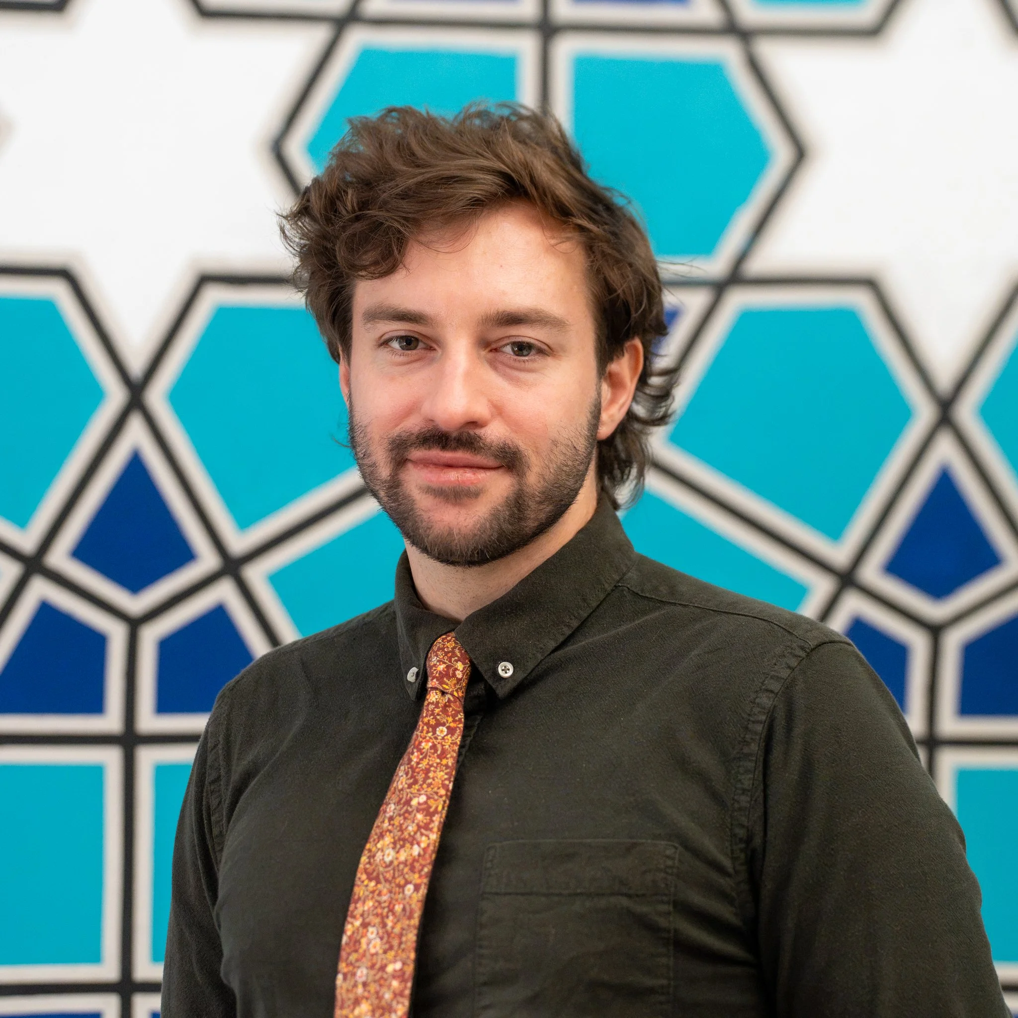 A young man with shoulder-length brown hair, a beard, and light skin, smiling, wearing a multicolored patterned sweater, standing against a plain white wall.
