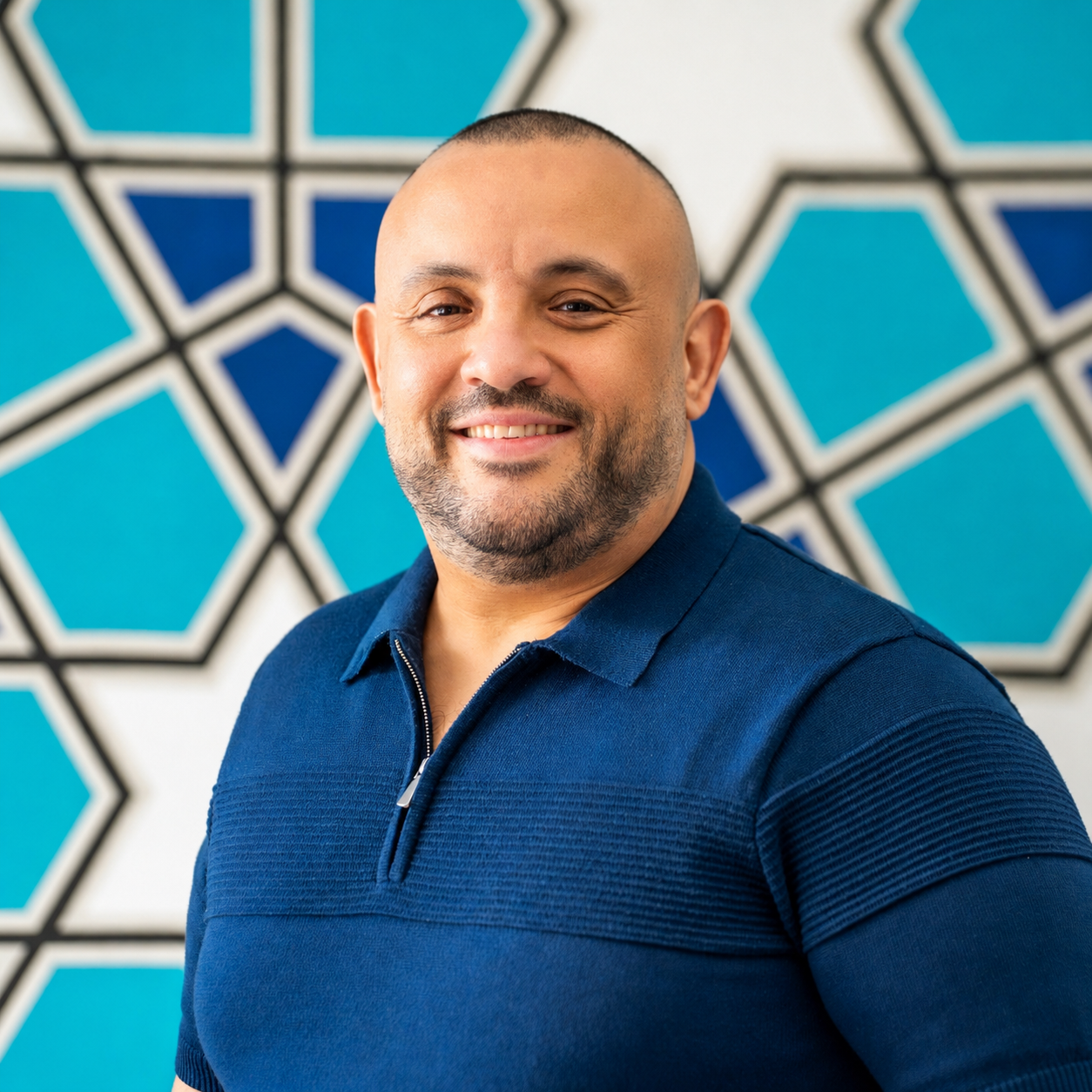 A man smiling, sitting at a table in a casual setting, wearing a black shirt and a blue jacket, with a blurred background.