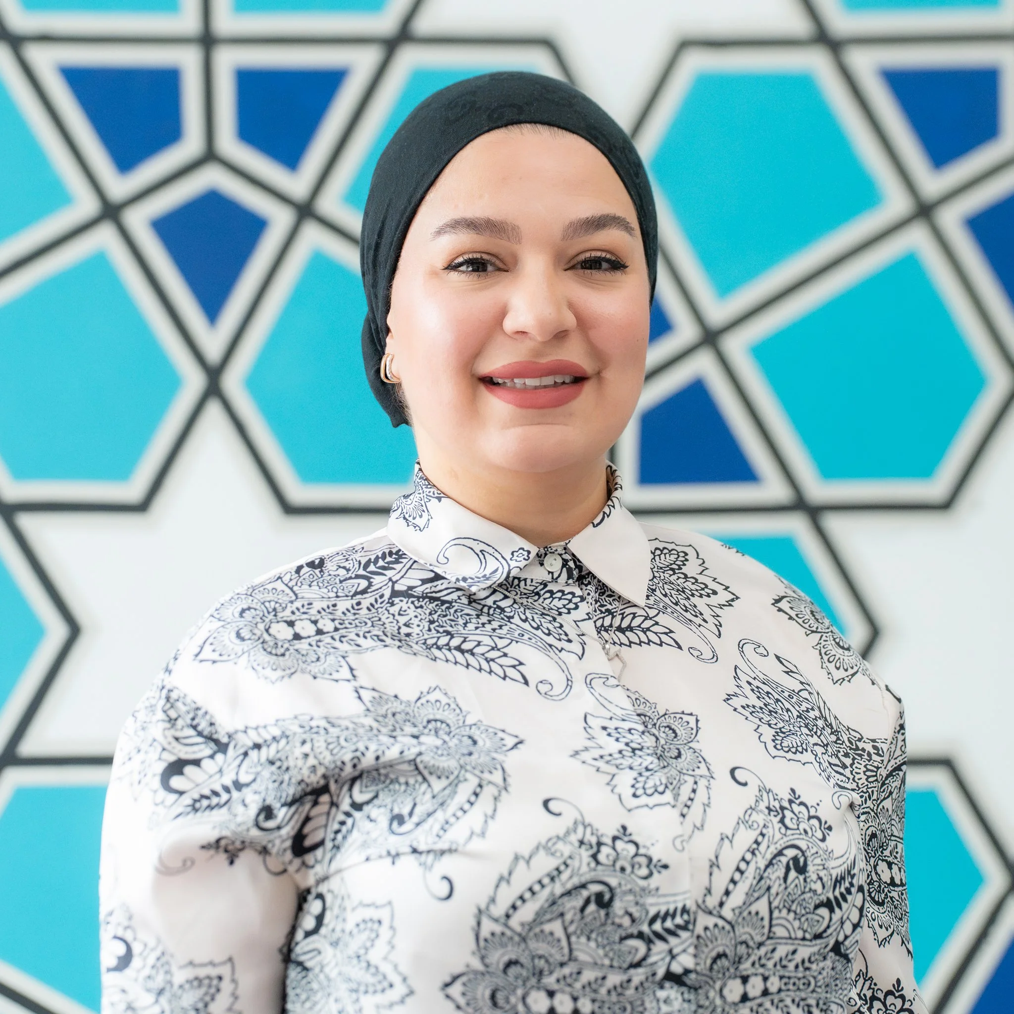 A smiling woman wearing a light-colored headscarf and a gray patterned blouse.