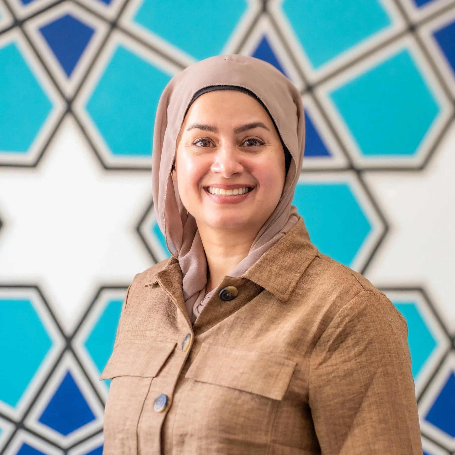 A woman with fair skin, dark hair, and brown eyes wearing a pink headscarf and navy blue top, standing against a plain light background.