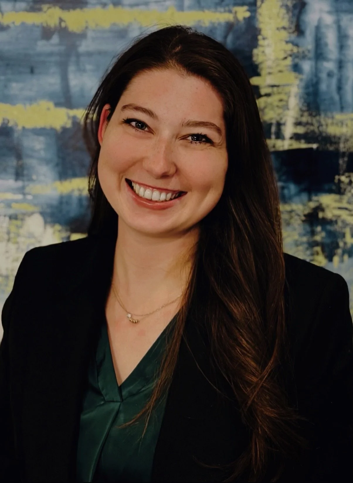 A woman with long dark hair smiling, wearing a dark blazer and green blouse, standing in front of an abstract blue and yellow background.