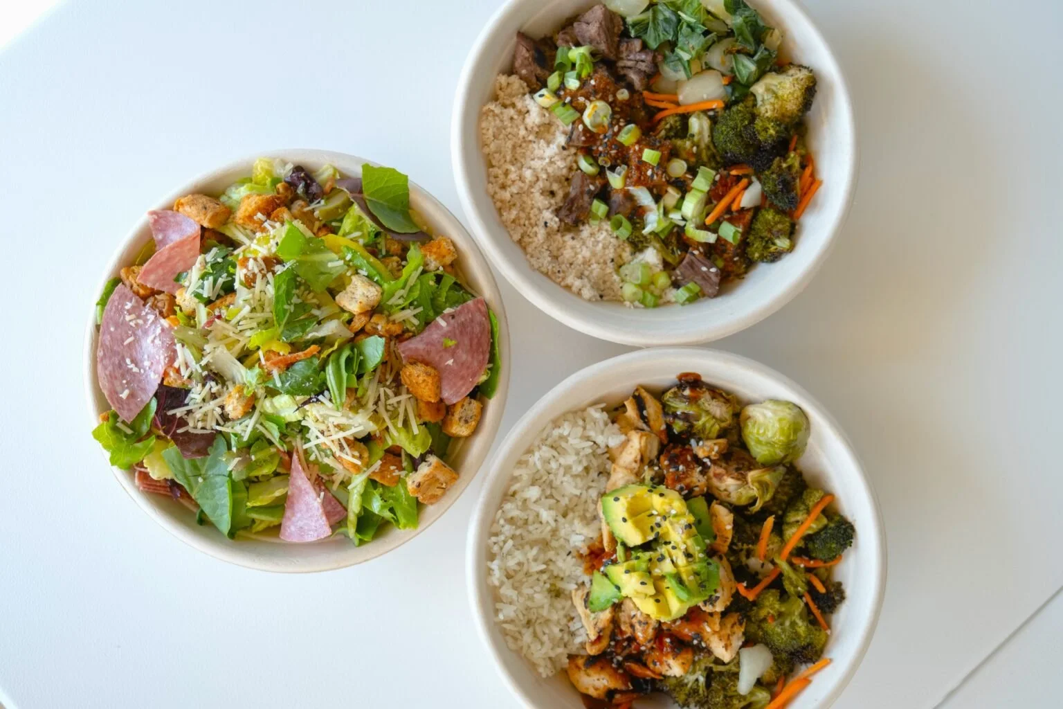 Three bowls of healthy food on a white surface. The left bowl contains a salad with mixed greens, diced chicken, shredded cheese, croutons, and sliced ham. The top right bowl has a grain base with roasted vegetables, beef, chopped green onions, and a sauce. The bottom right bowl contains white rice topped with grilled chicken, sliced avocado, vegetables, and a dark sauce.