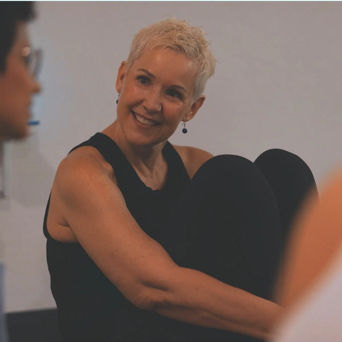 A woman with short blonde hair smiling and looking at a person with glasses in a conversation.