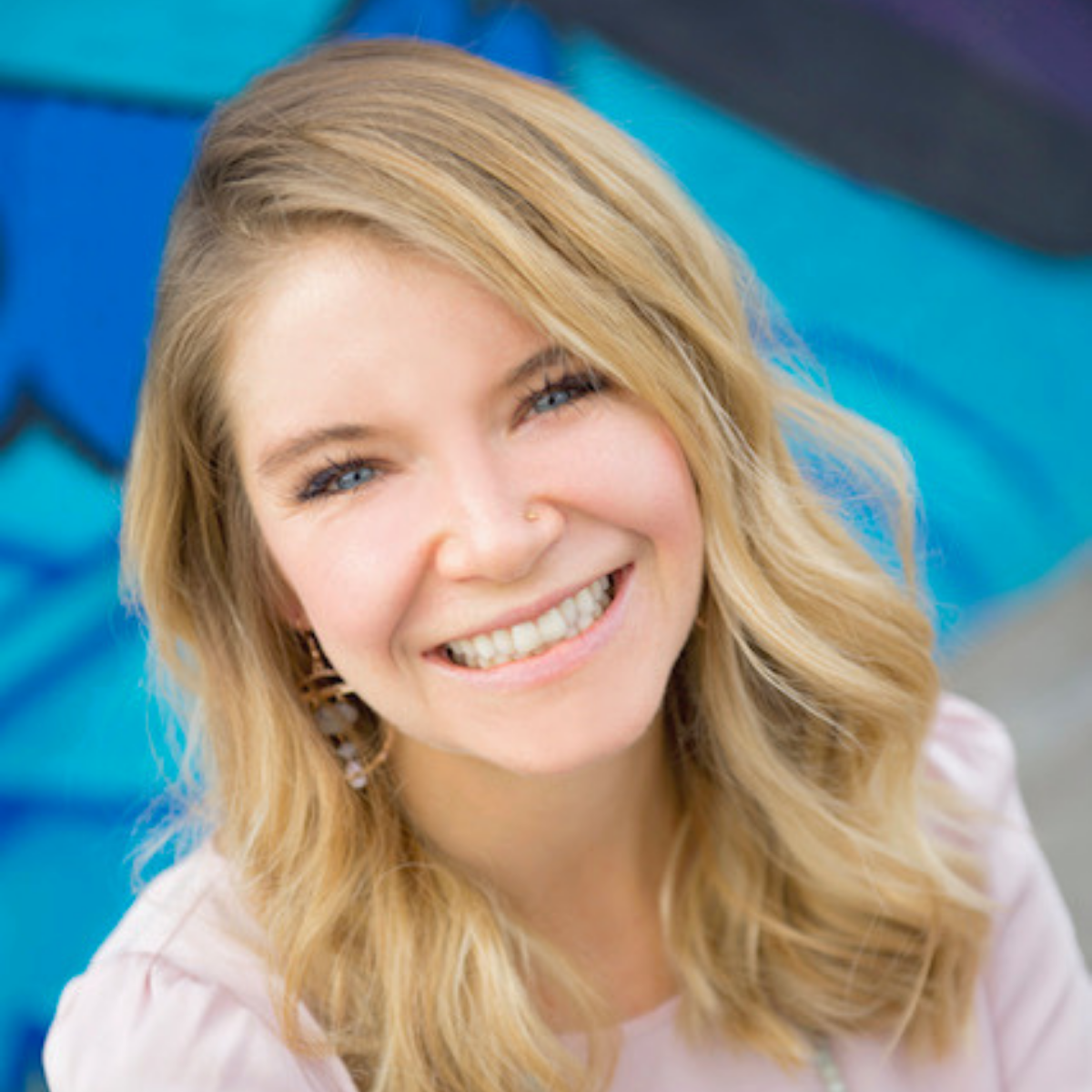 Close-up of a smiling woman with blonde hair, wearing earrings, in front of a blue background.