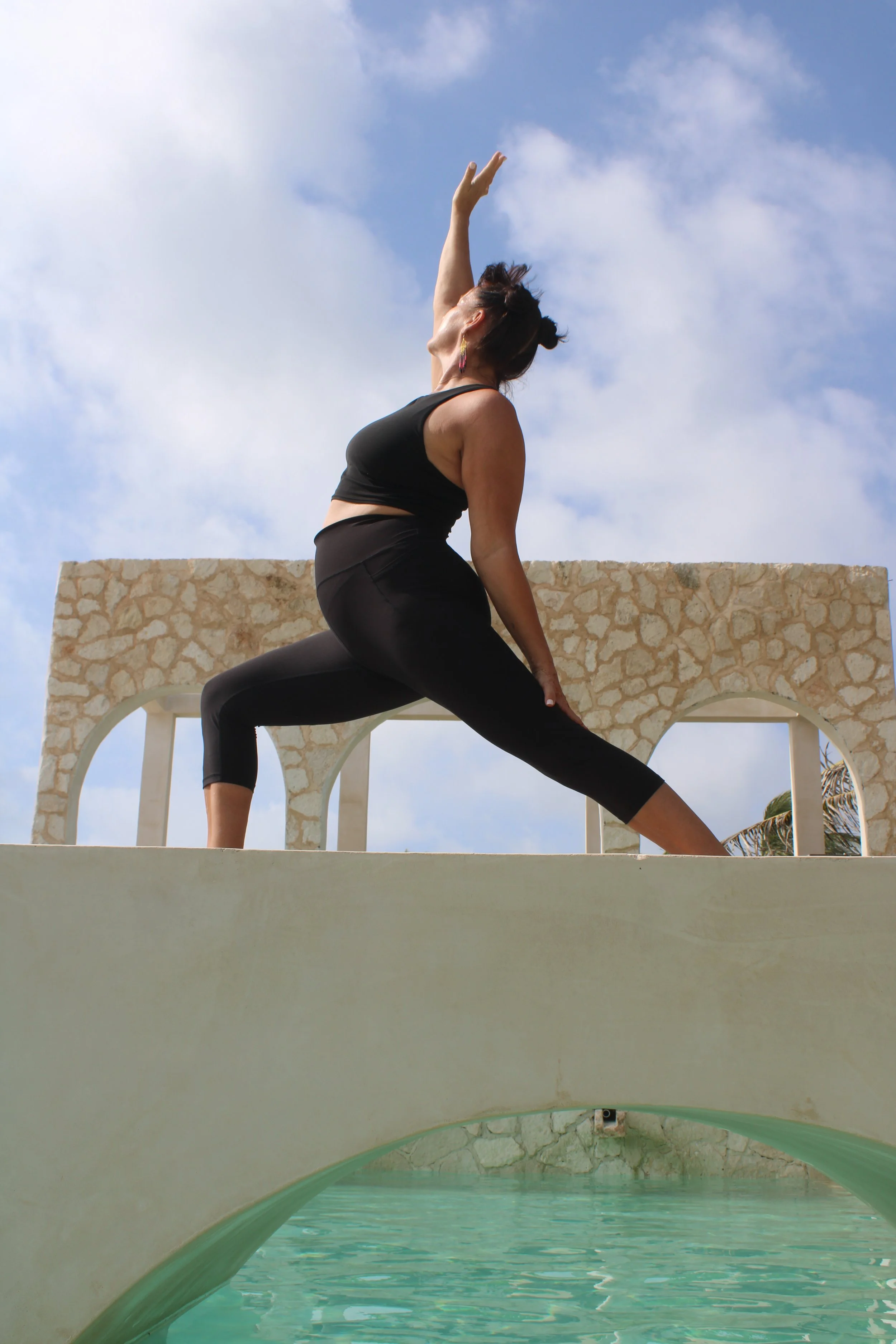 Woman practicing yoga on a bridge over a pool outdoors, with a stone wall and cloudy sky in the background.