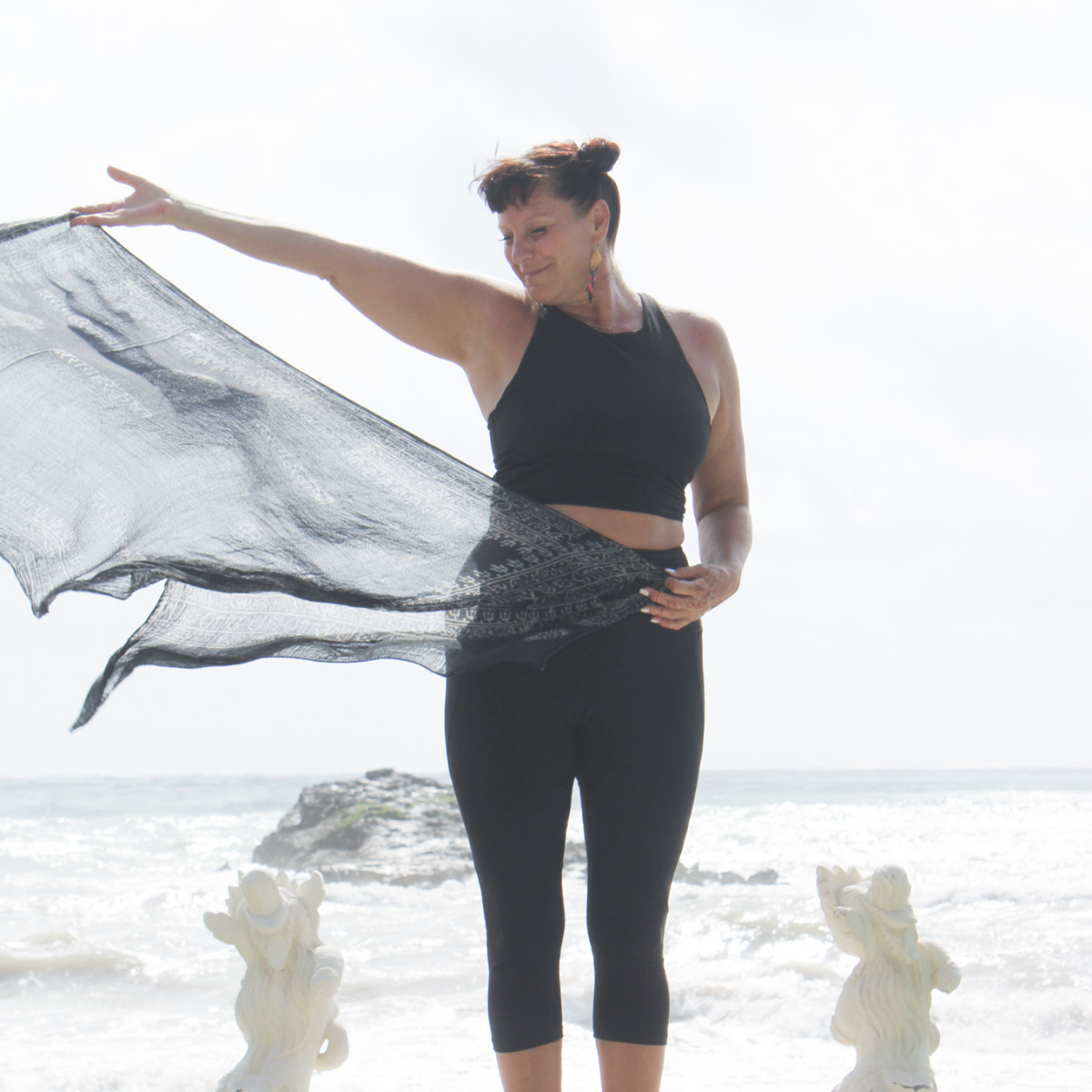 A woman in black yoga attire standing on a beach, holding a sheer black fabric, with two small gorilla statues at her feet, and ocean waves in the background.