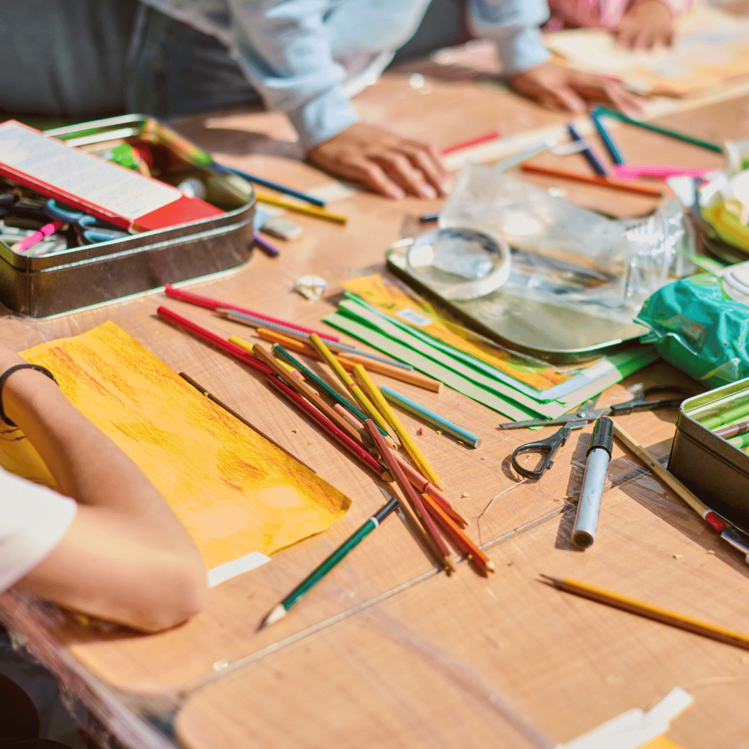 art supplies on table with arms and hands of three people around table