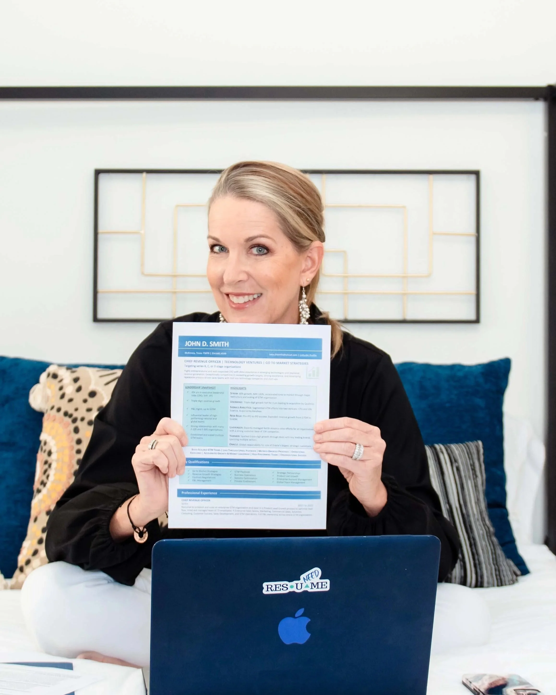 A woman with blonde hair, wearing earrings and a black jacket, smiling and holding a resume in front of her. She is sitting at a desk with a blue Apple laptop in front of her that has a sticker that says "RESU ME NEED" on it. Behind her is a framed artwork with horizontal gold and black lines on a white background, and a plush leopard print pillow sitting on a chair to her left.