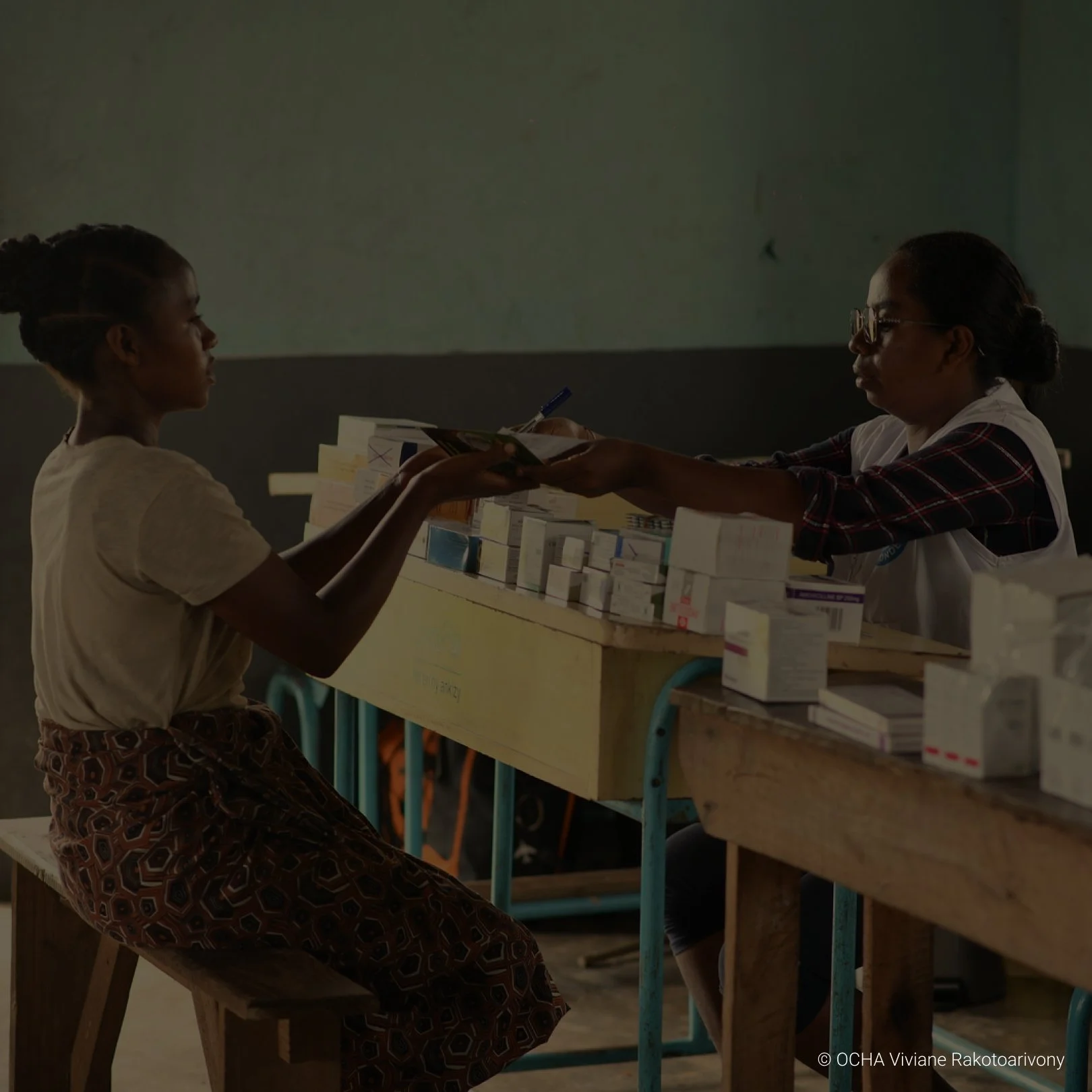 A young girl sitting on a wooden bench, receiving medication from a woman at a table with various boxes of medicine, in a dimly lit room.