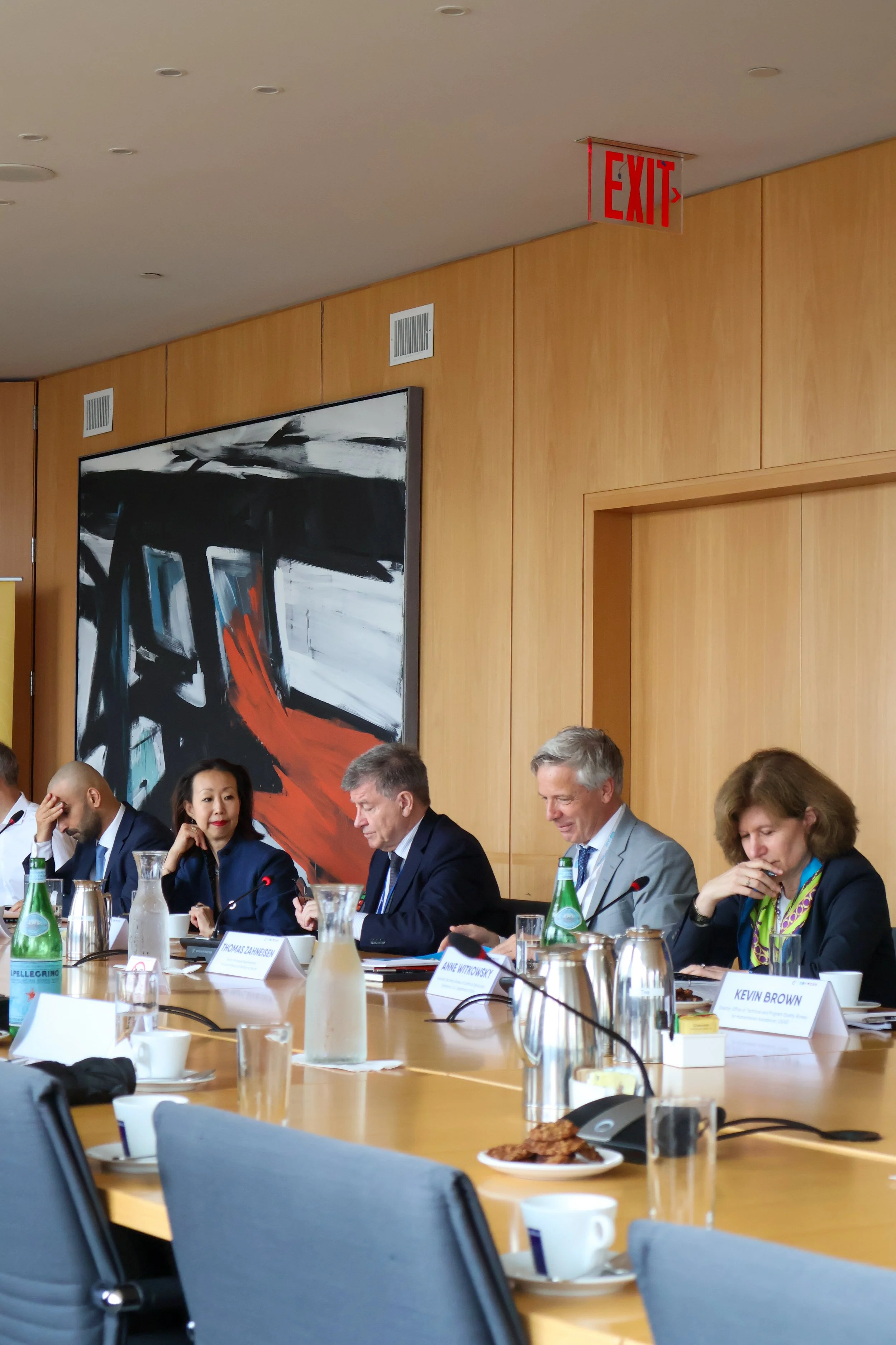 Business meeting with six people seated at a conference table, several bottles of water, glasses, plates with food, and nameplates in front of each person, with a large abstract painting on the wooden wall behind them.