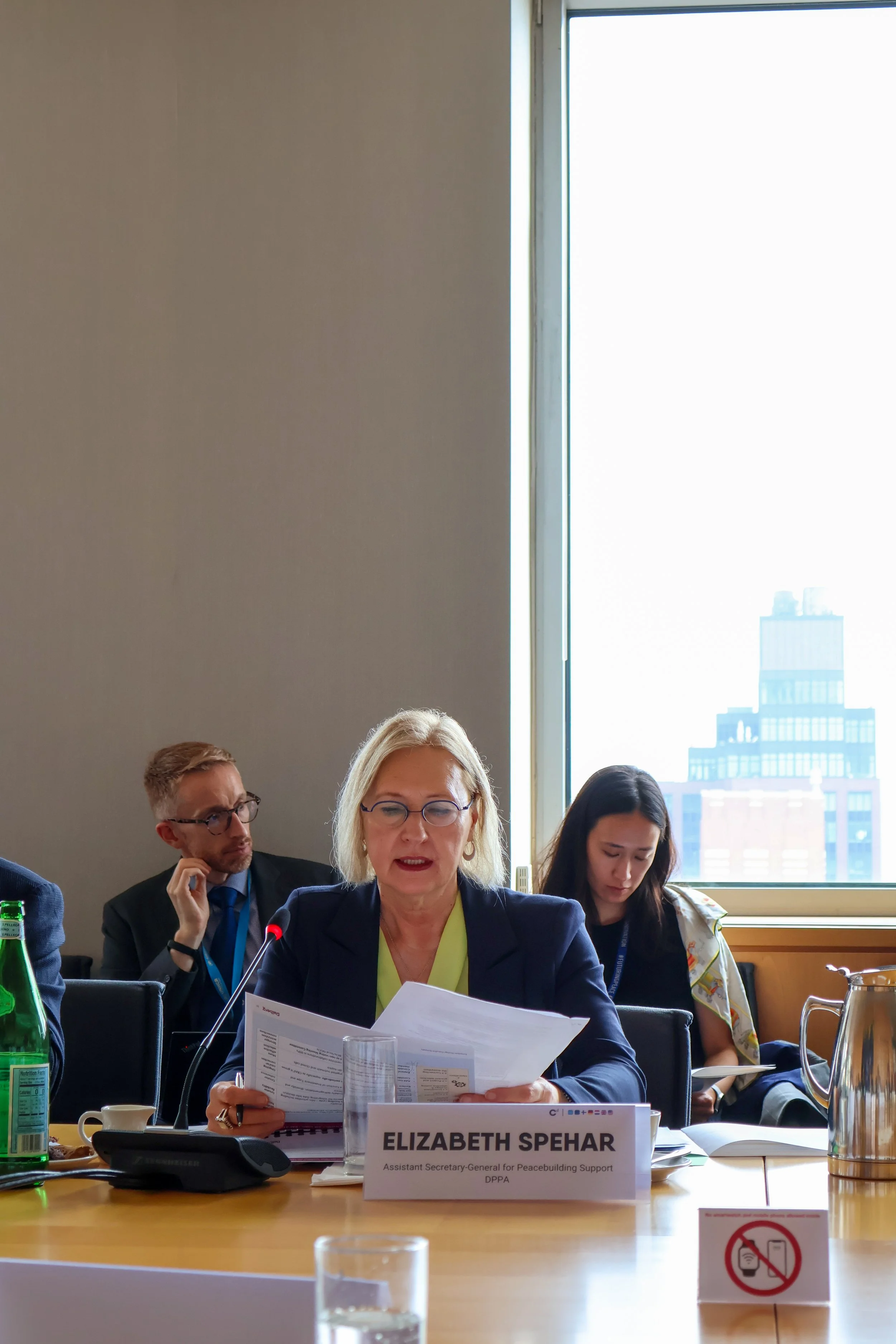 Elizabeth Spehar sitting at a conference table, reading documents, with a nameplate indicating her as Assistant Secretary-General for Peacebuilding Support. Other attendees are visible in the background.