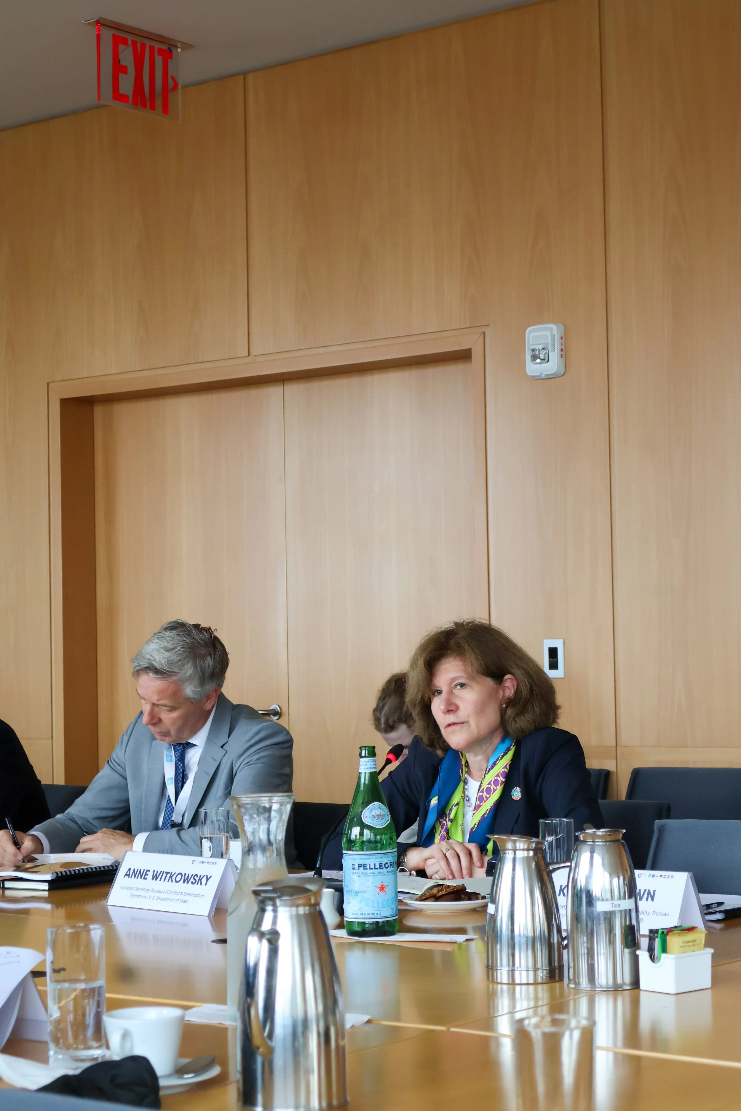 Women and men sitting at a conference table during a meeting, with nameplates, water bottles, and coffee cups. A woman in a dark jacket and a scarf is looking ahead, while a man in a gray suit is writing.