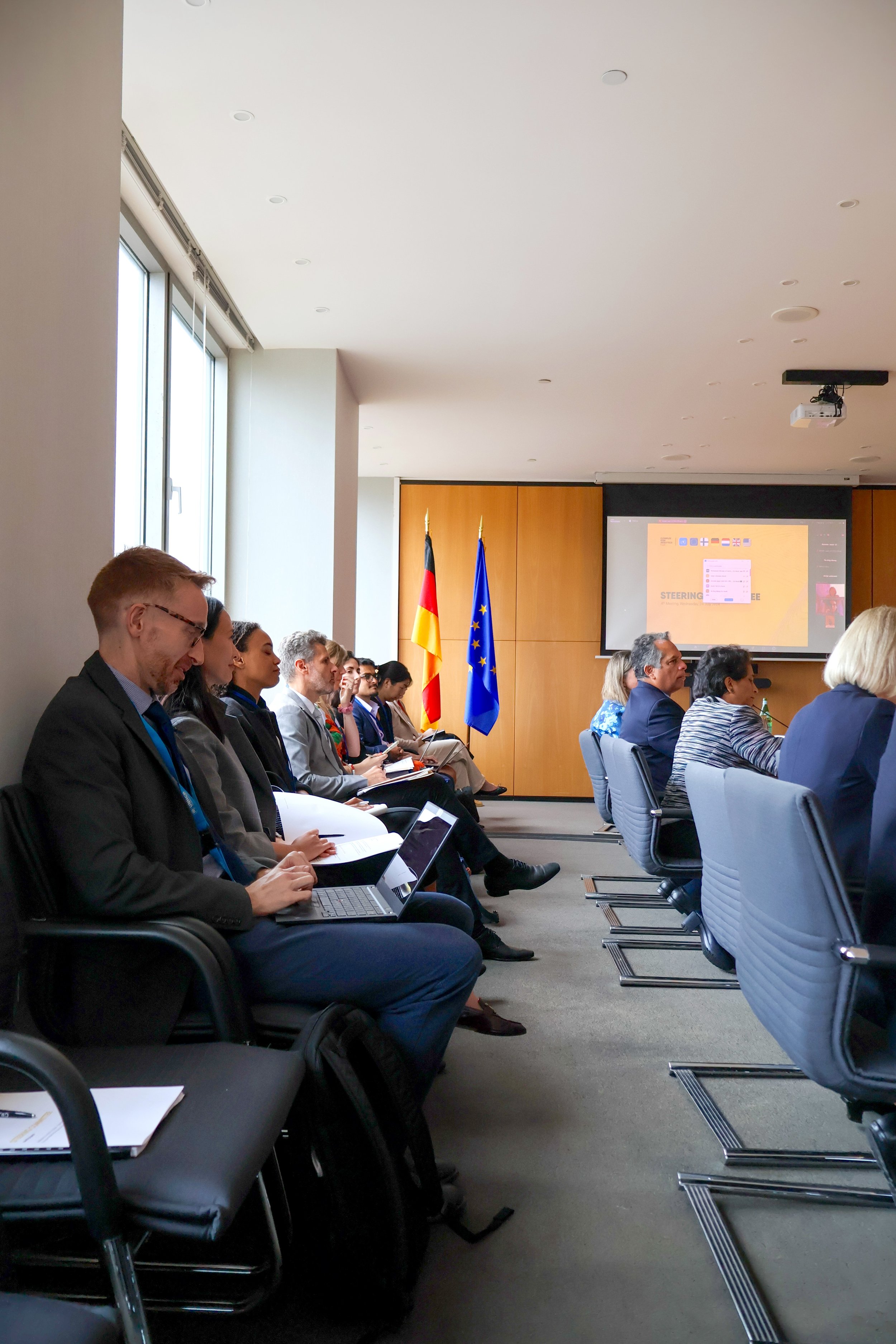 Attendees in business attire sitting in a conference room during a meeting, with flags of Germany and the European Union in the background, and a presentation projected on the wall.