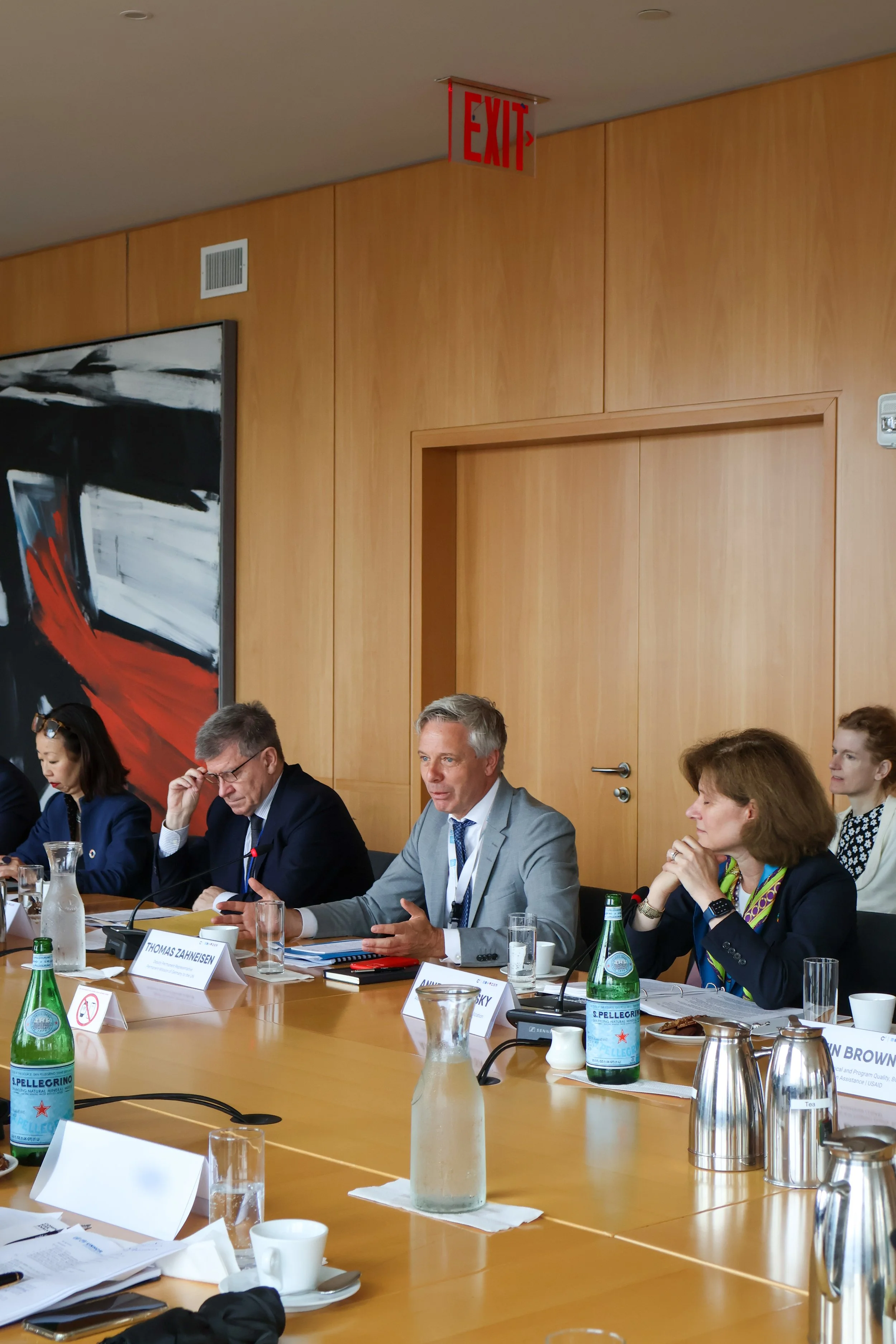 A group of six people sitting at a conference table, engaged in a discussion. The table has drinks, notebooks, and nameplates. The room has wooden walls, a large abstract painting, and a red "EXIT" sign hanging from the ceiling.
