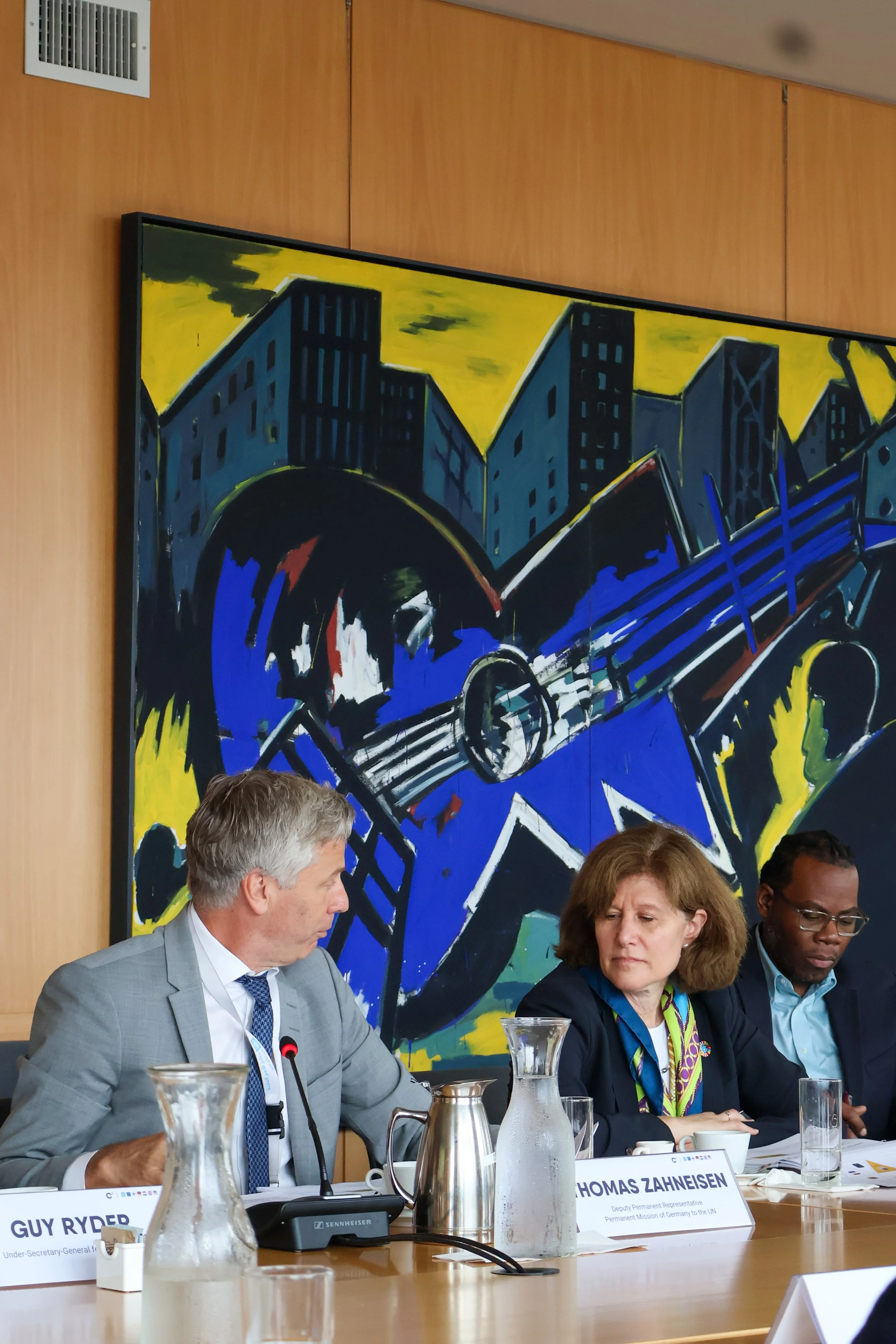 Three people seated at a conference table in front of a large colorful abstract painting of a cityscape, with nameplates, glasses, and pitchers on the table.