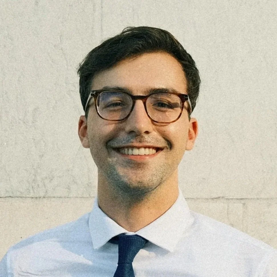 A young man wearing glasses, a white collared shirt, and a dark tie, smiling with a plain wall background.