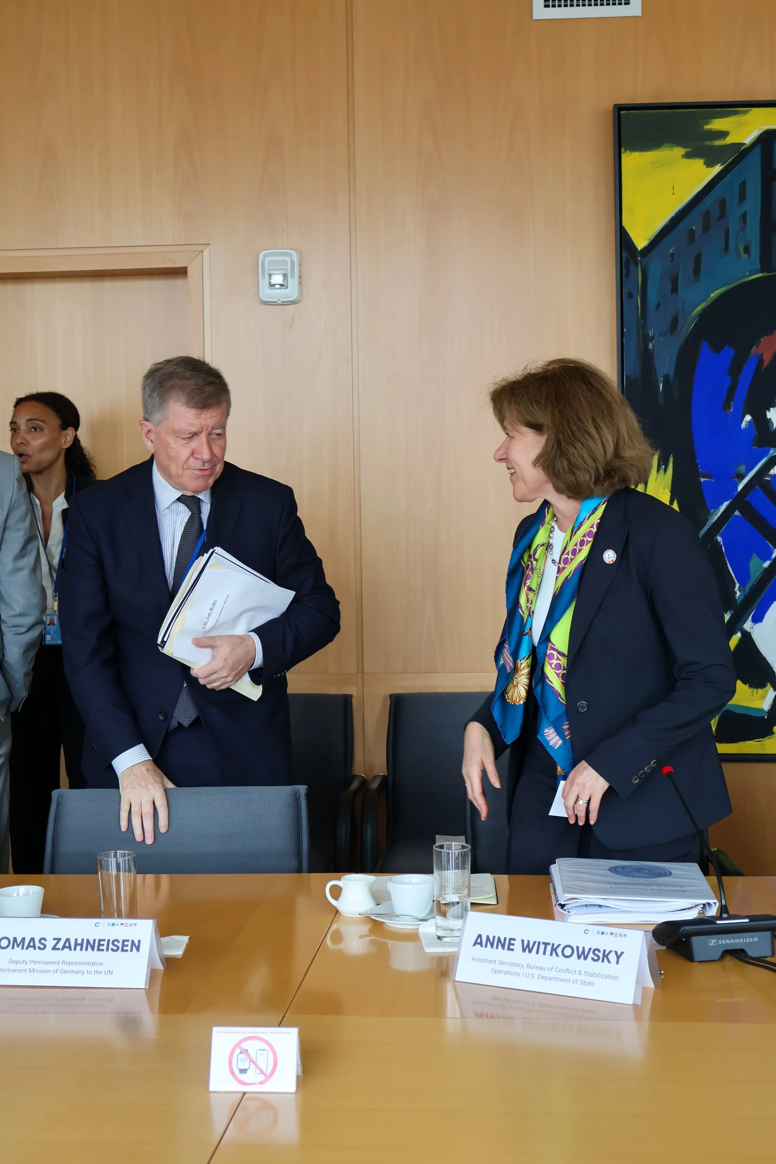 Two women and a man engaged in conversation at a conference table. The woman on the right is smiling, wearing a dark blazer and colorful scarf. The man on the left is holding documents and wearing a dark suit. There are nameplates for Tomas Zahnisen 