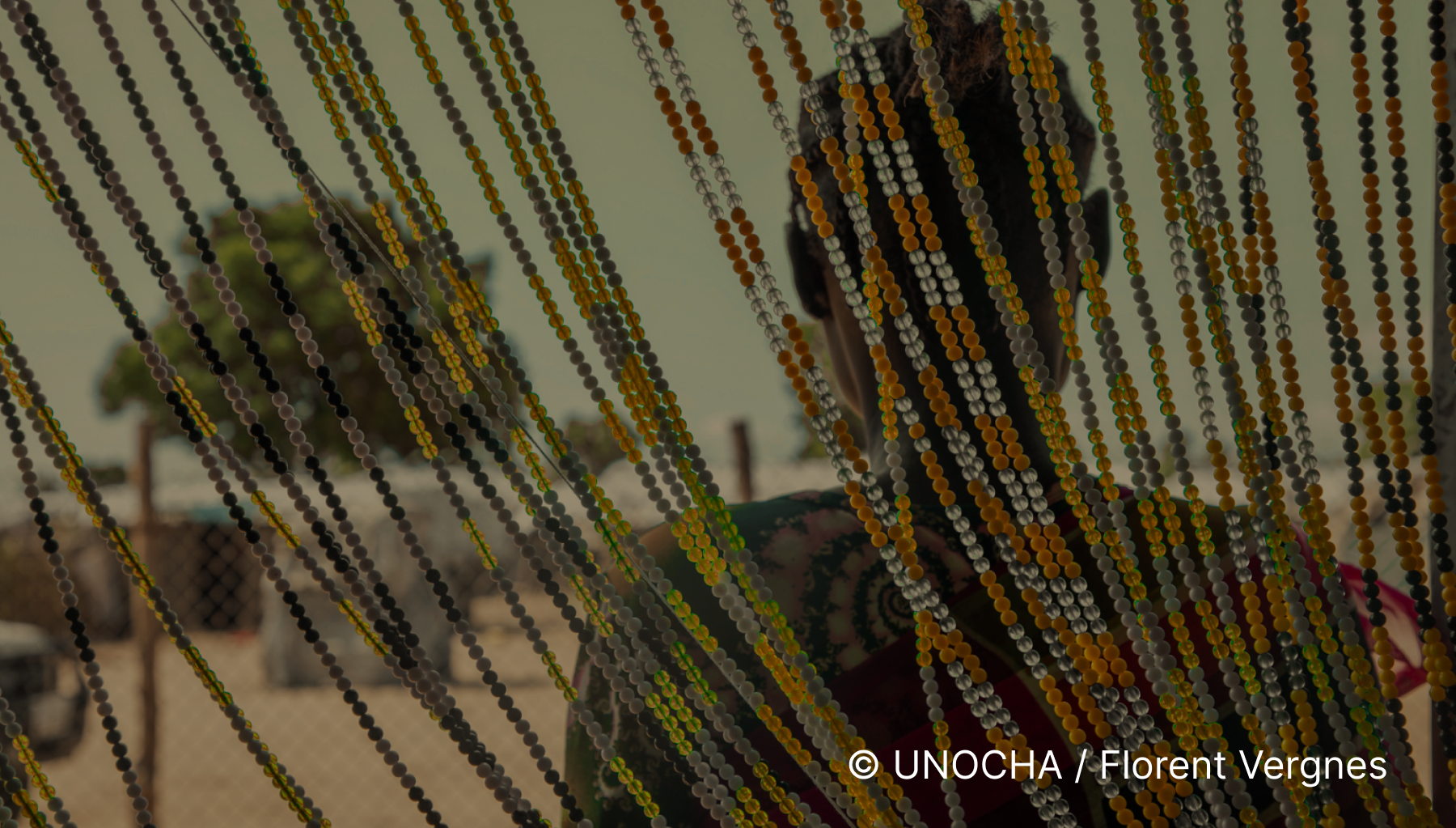 A person behind a beaded curtain made of multicolored beads, with trees and a fence visible in the background.