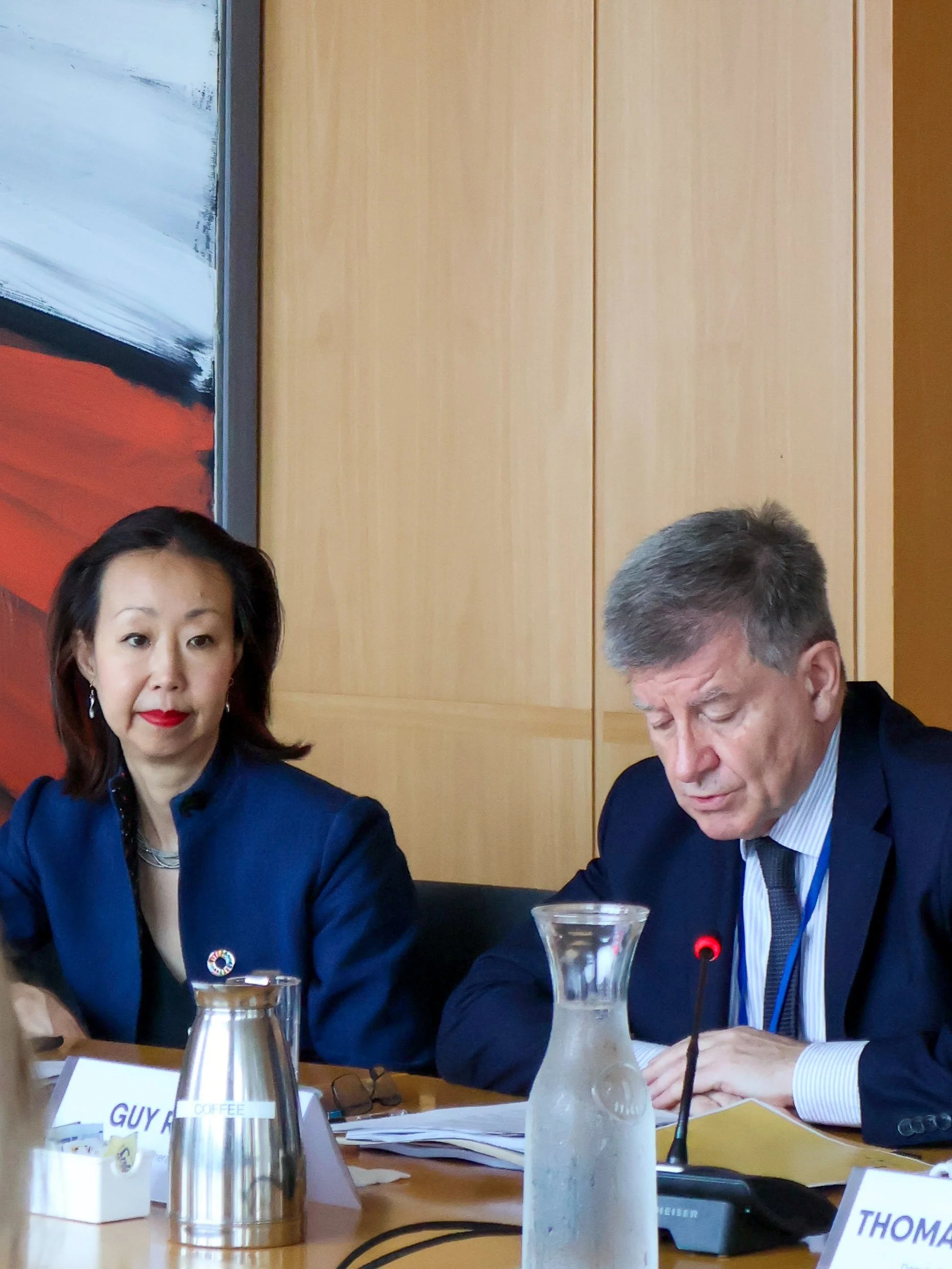 Two people seated at a meeting table with documents, a microphone, and water pitchers.