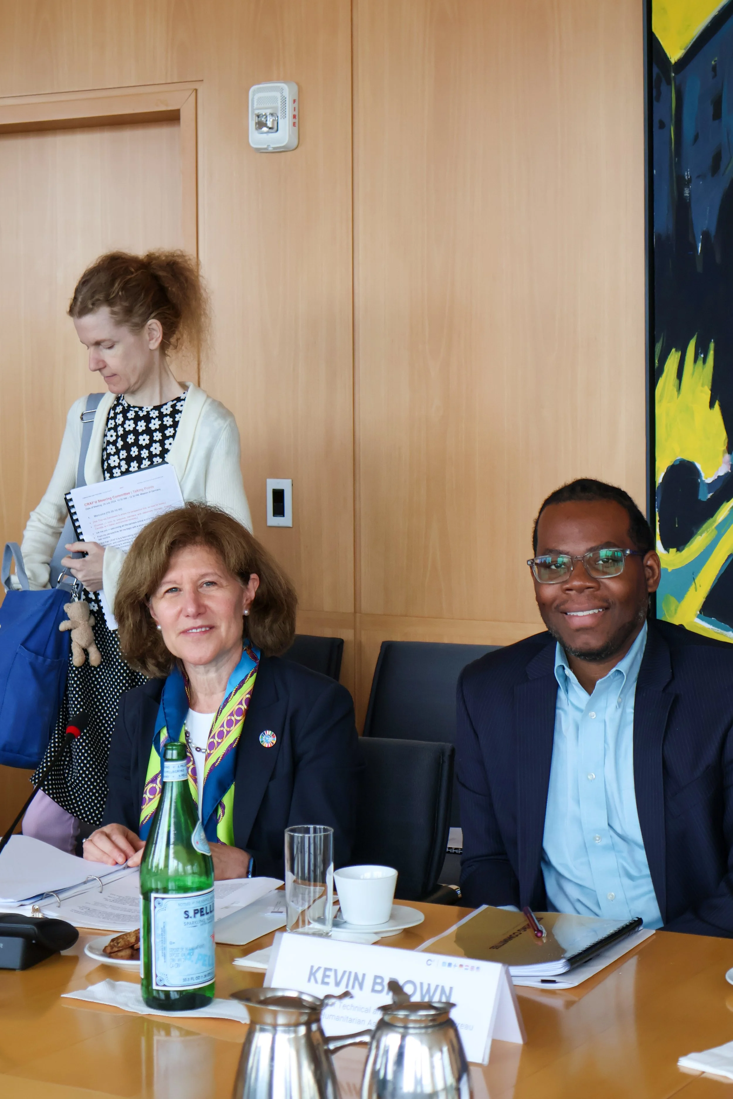 Two women and one man sitting at a conference table with documents and drinks, one woman standing with a notebook.