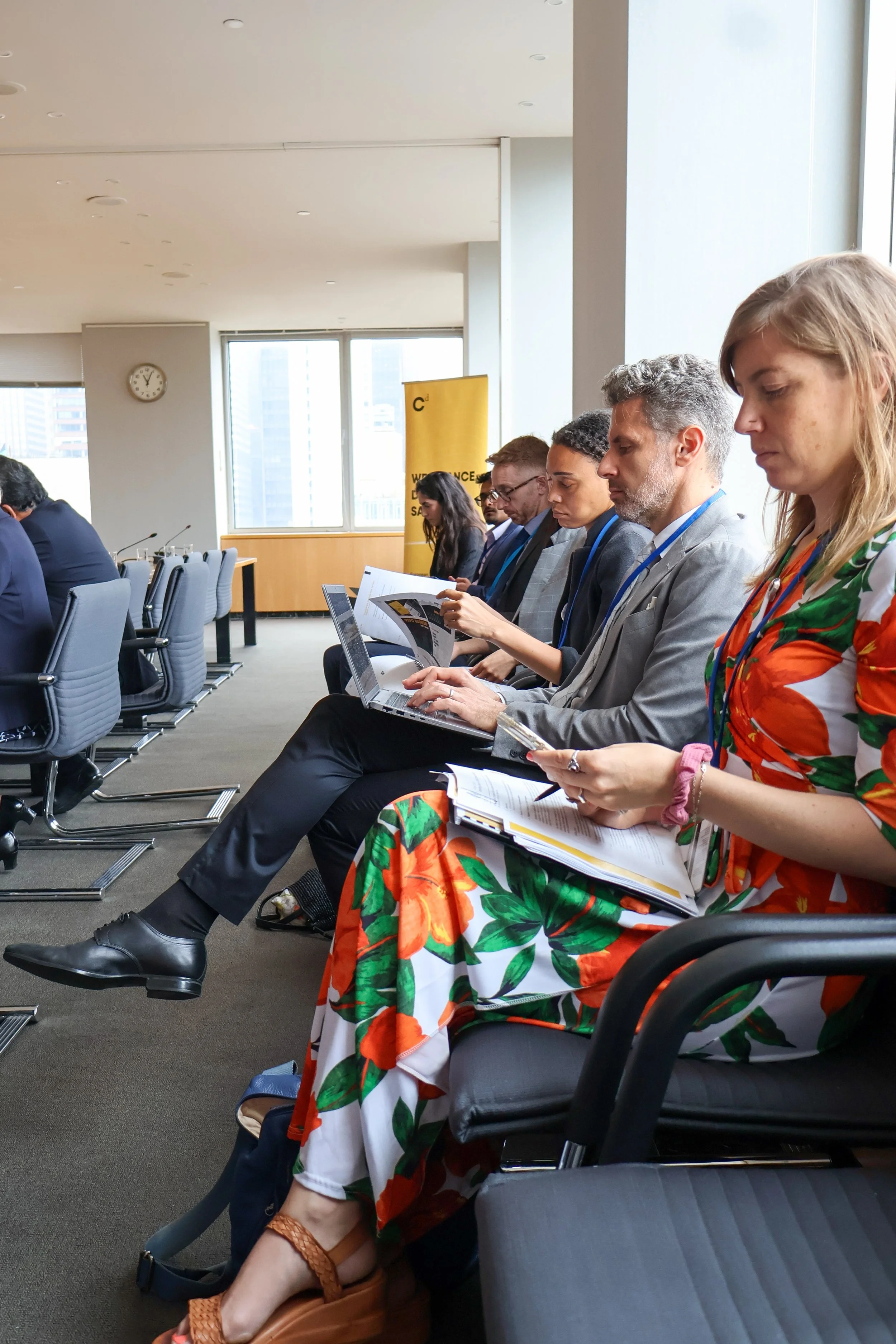 A group of seven business professionals sitting in a row at a conference, working on laptops and reviewing documents. They are in a modern office with large windows and city buildings in the background.