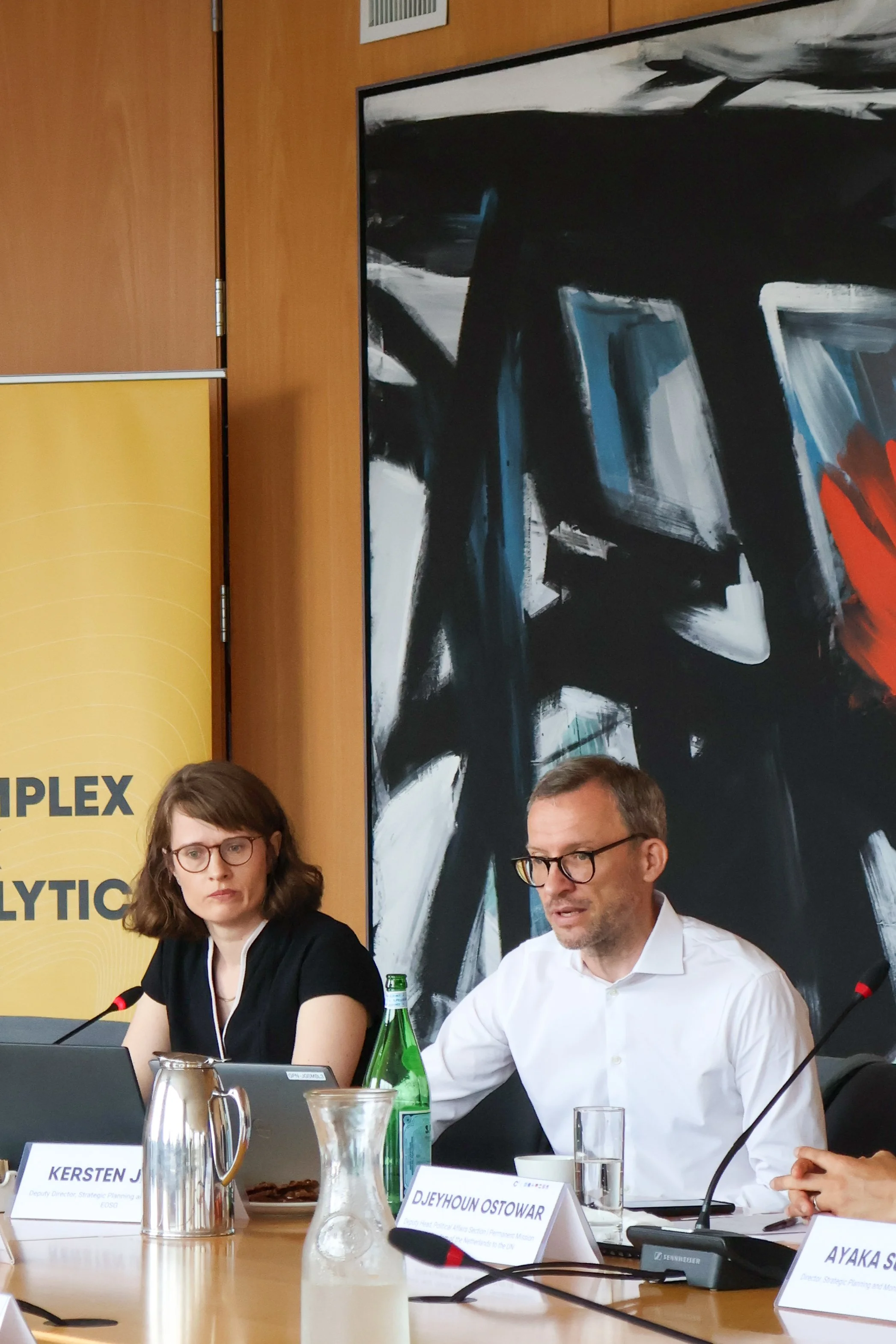 Two individuals seated at a conference table with microphones, nameplates, and drinks, participating in a formal discussion or panel. There is a large abstract black and white painting on the wall behind them.